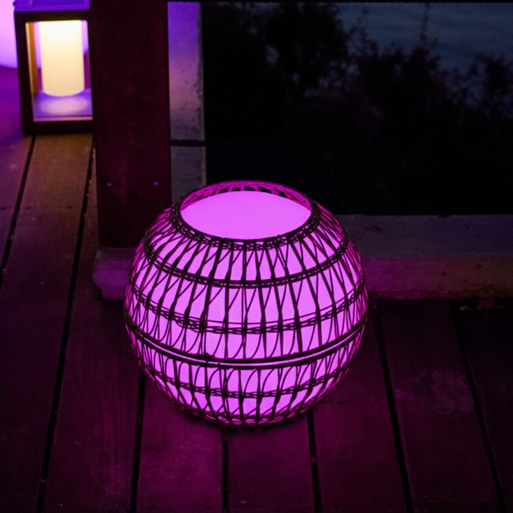 Eine runde, korbähnliche Lampe leuchtet pink auf einer Holzterrasse.