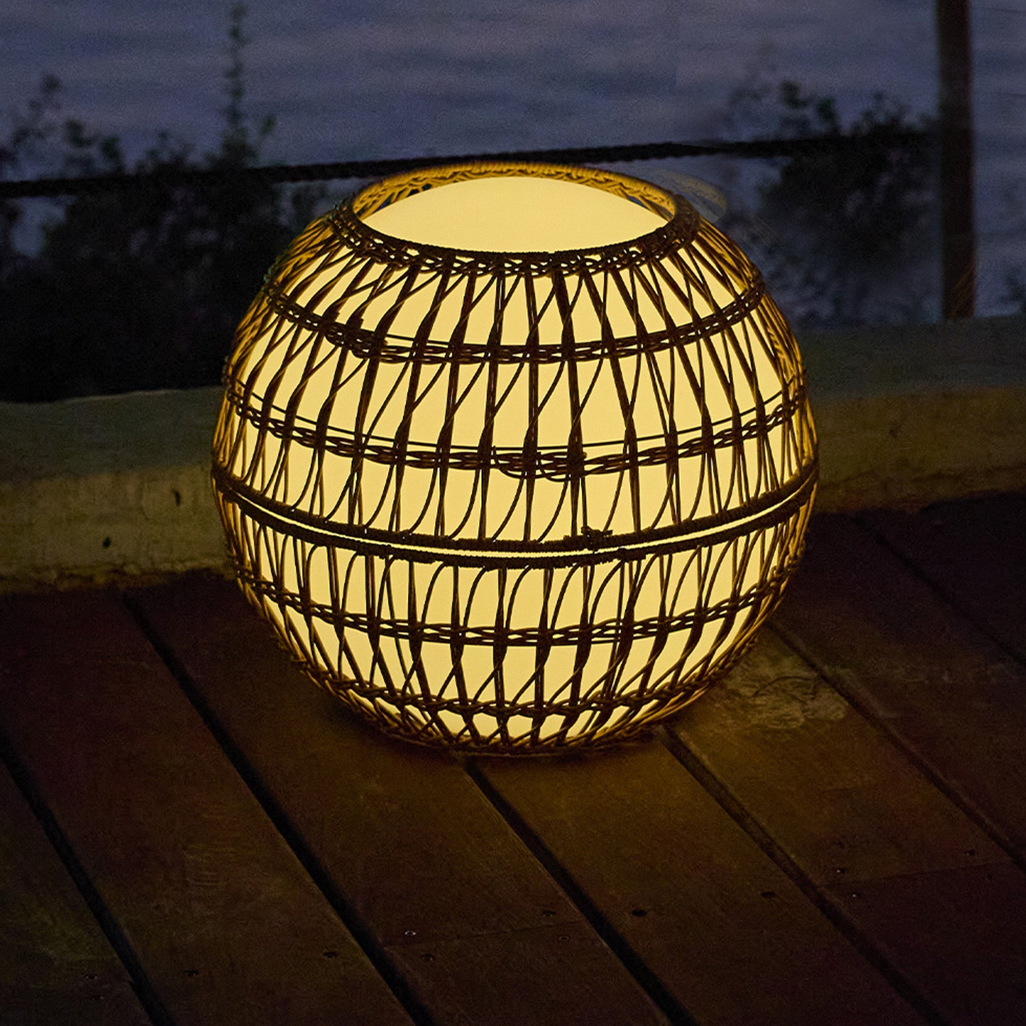Eine runde, geflochtene Lampe leuchtet gelb auf einer Holzterrasse im Freien bei Dämmerung.