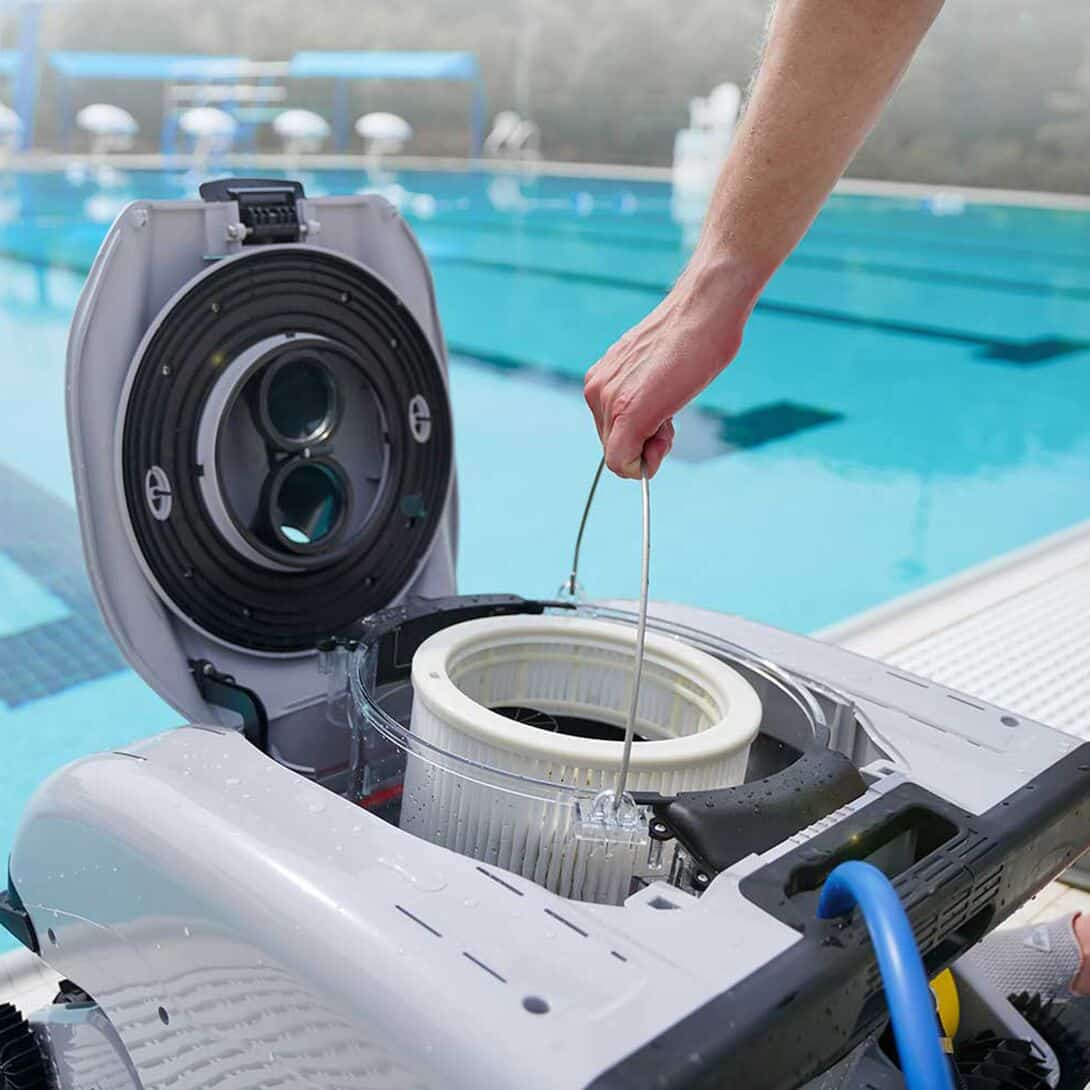 Una mano levanta un robot de piscina con la tapa abierta y el filtro expuesto. Piscina al fondo.