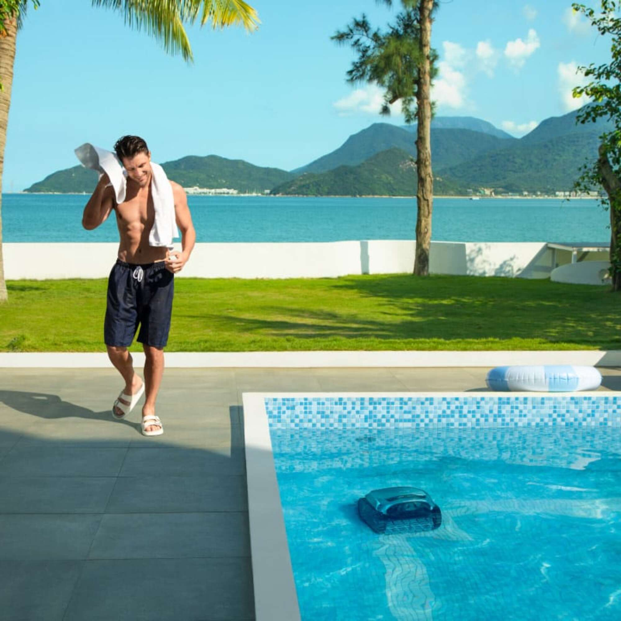 Mann mit Handtuch im Poolbereich, Meer und Berge im Hintergrund.