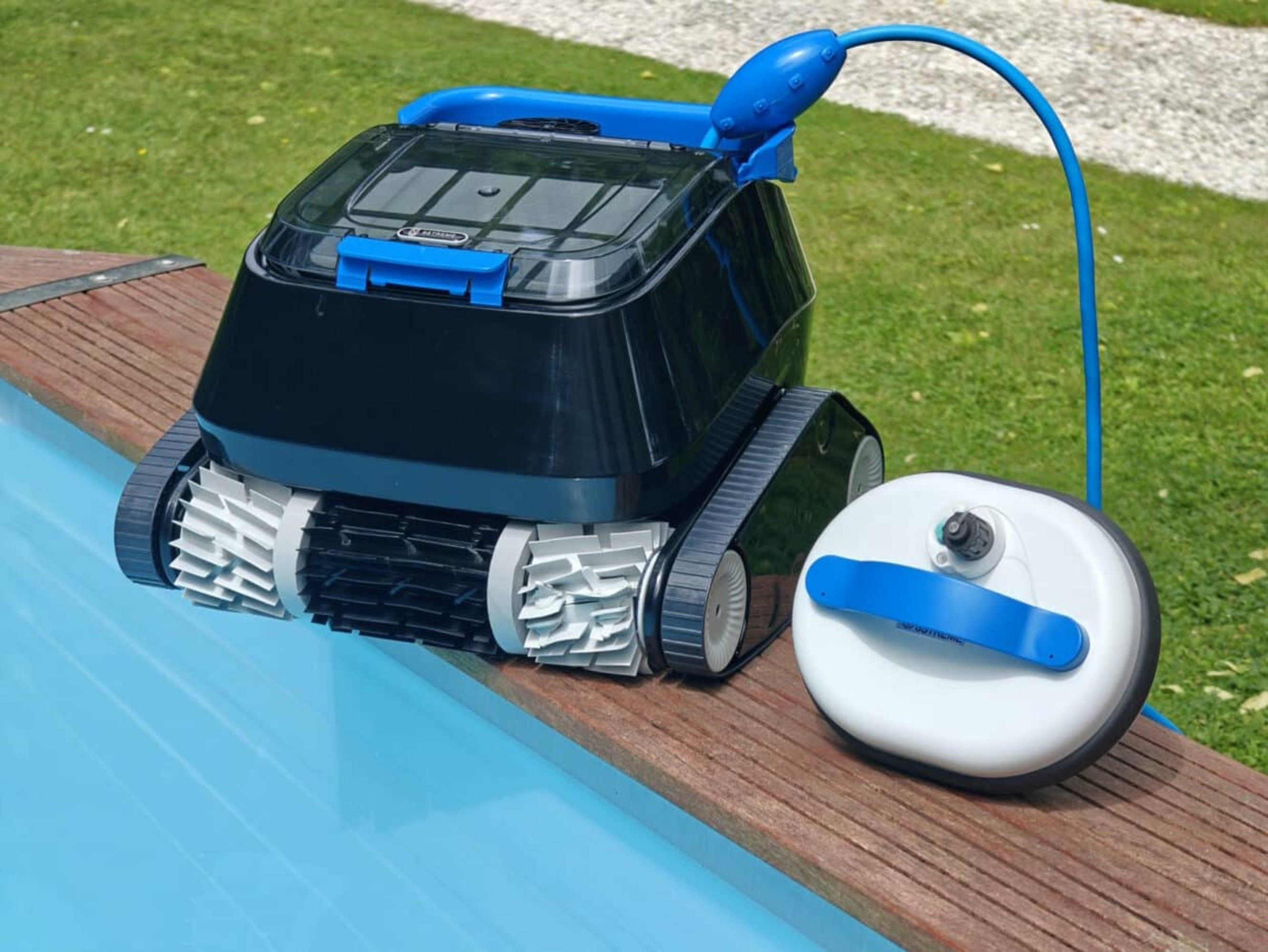 Un robot de piscina negro con mango y manguera azules está sobre una cubierta de madera junto a una piscina.