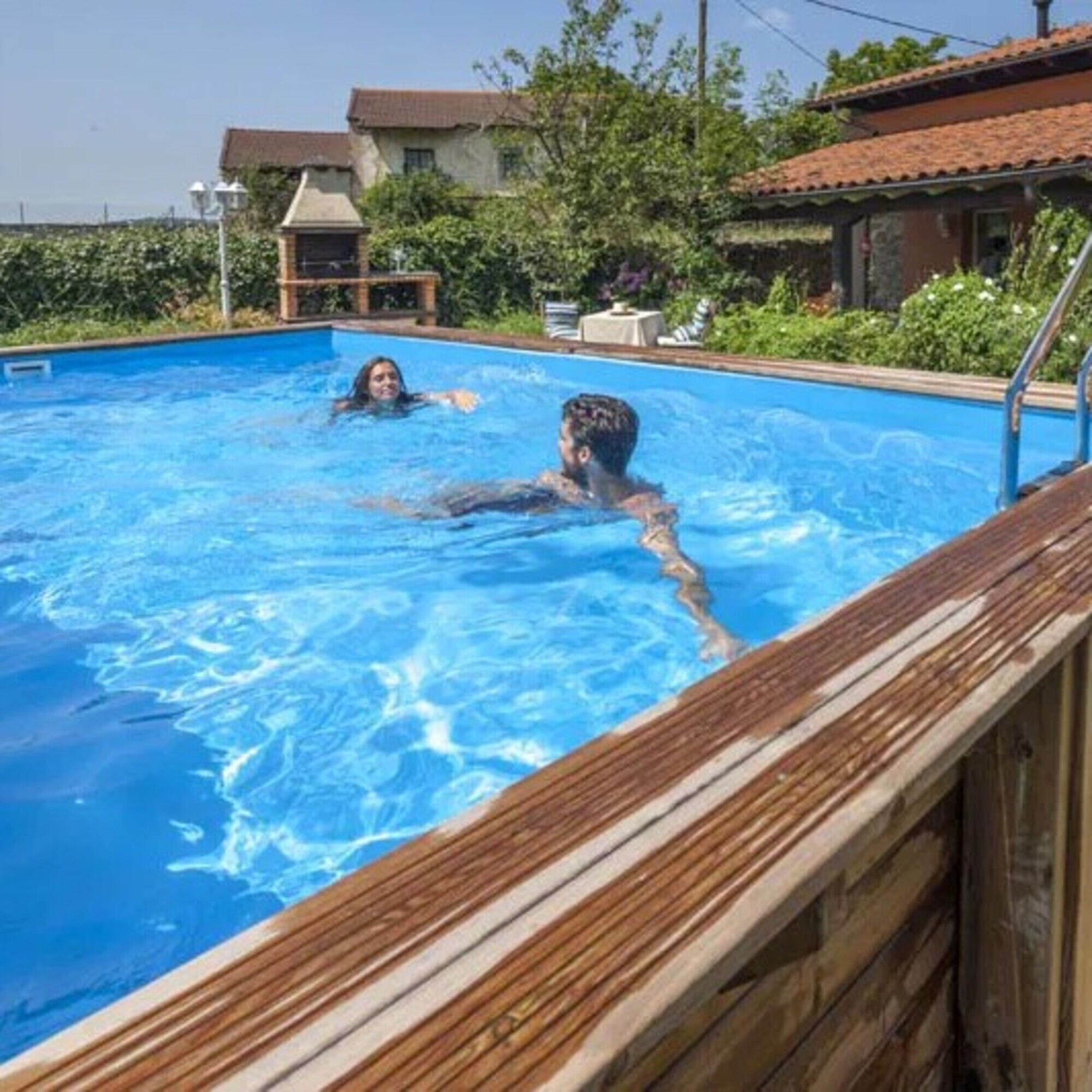 Una pareja nada en una piscina azul en un día soleado, casas al fondo.