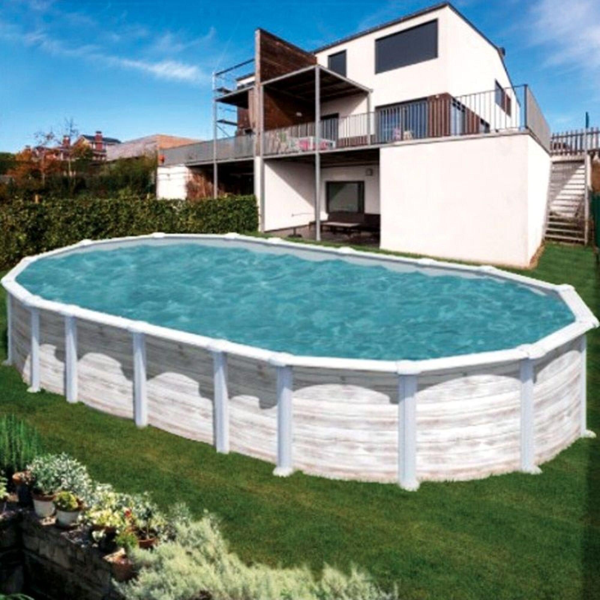 Una piscina ovalada con agua azul se encuentra en un jardín junto a una casa blanca.