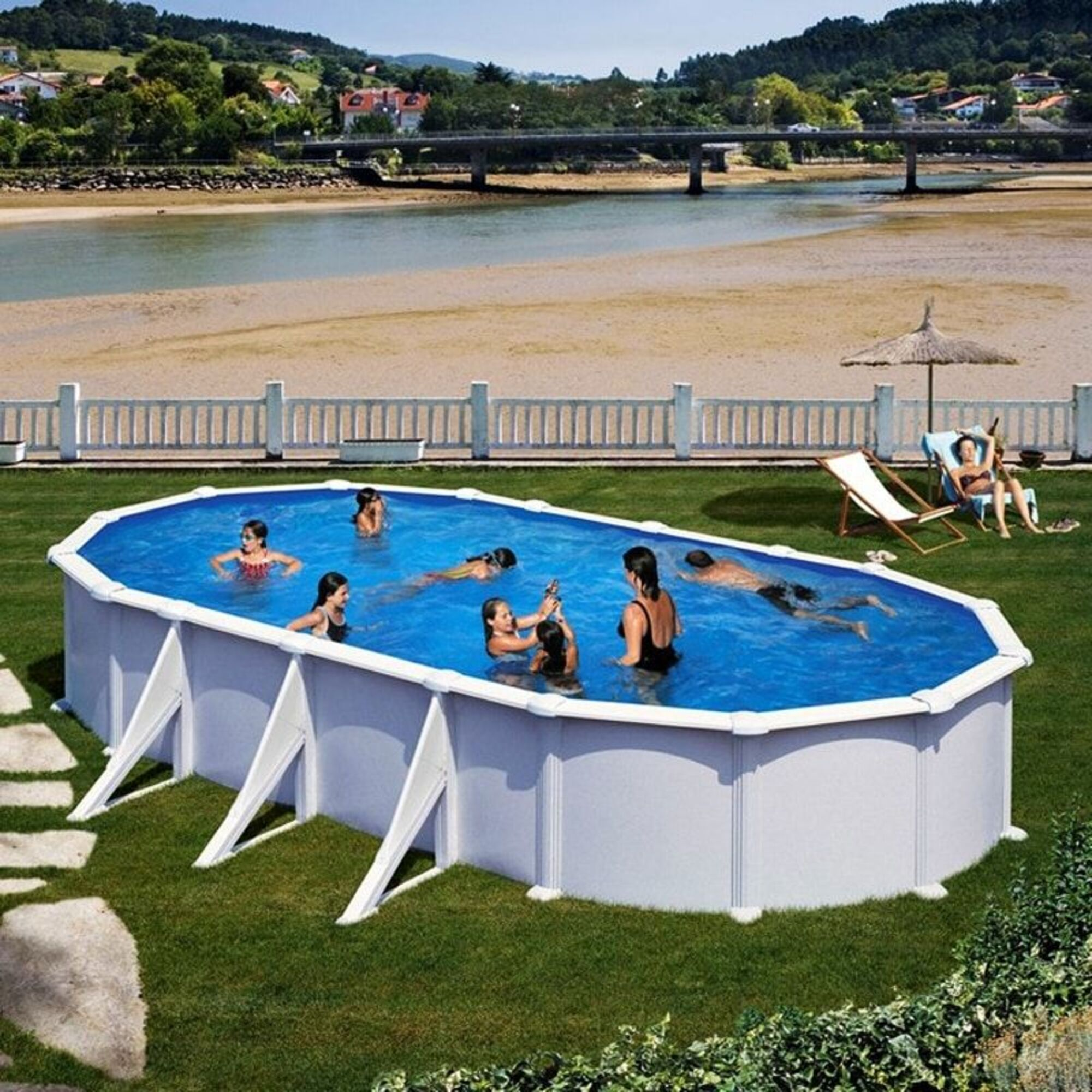 Personas nadan en una piscina ovalada en el césped, con un puente al fondo.