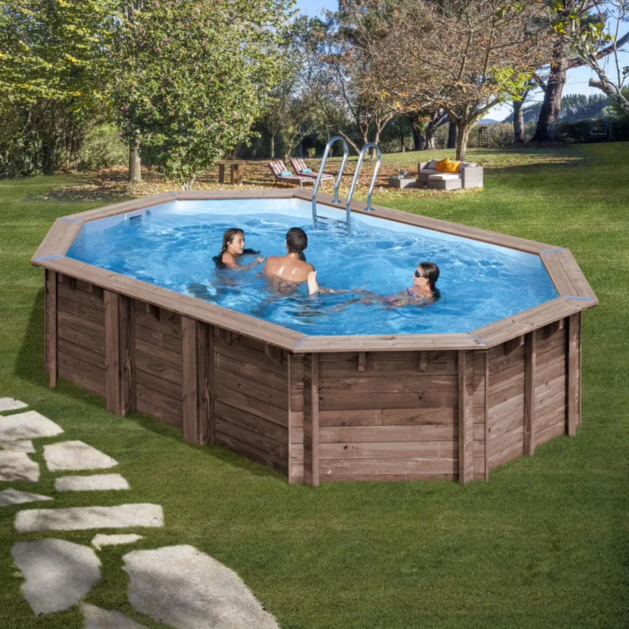 Tres personas nadan en una gran piscina octogonal de madera sobre el suelo en un patio trasero con césped y árboles.