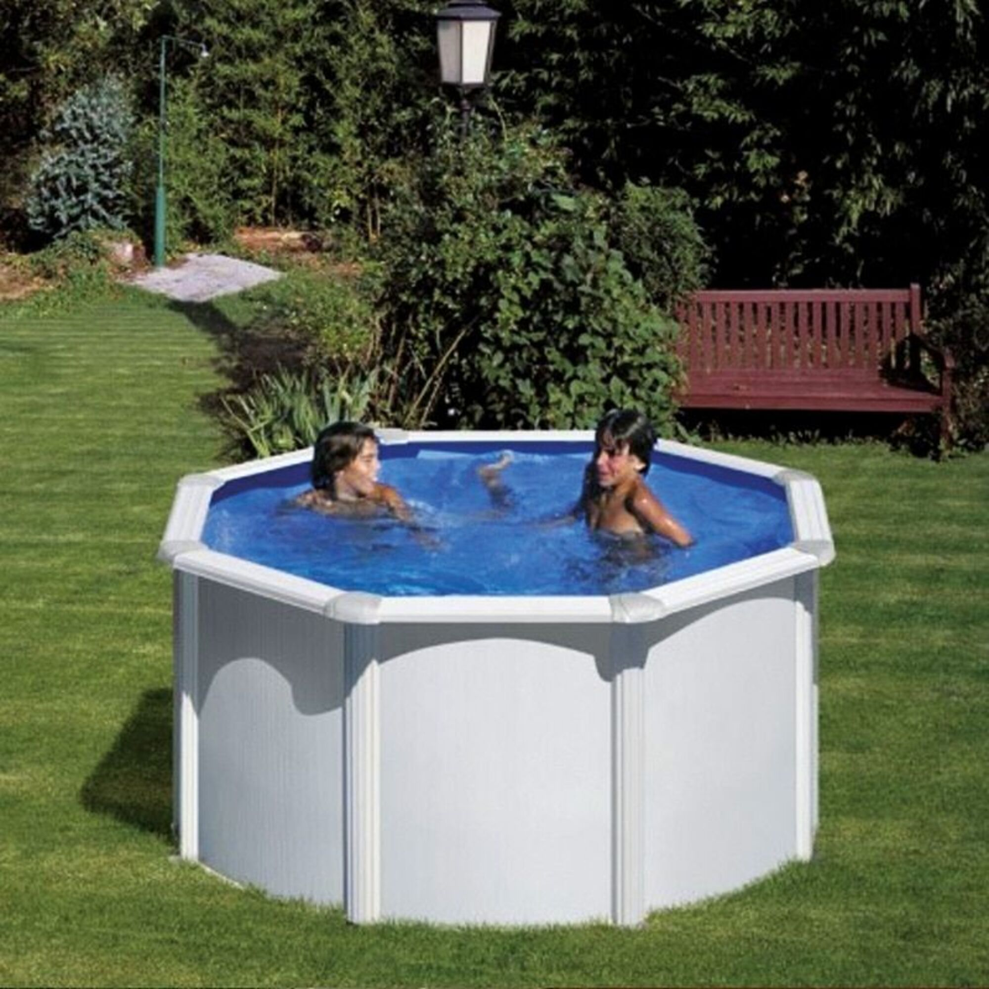 Dos niños chapotean en una piscina octogonal en un jardín. Un banco de madera al fondo.