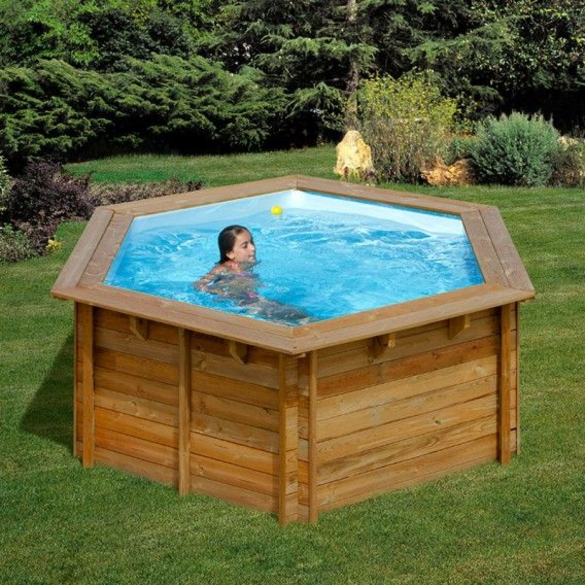 Una niña nada en una piscina hexagonal de madera en un jardín con árboles al fondo.