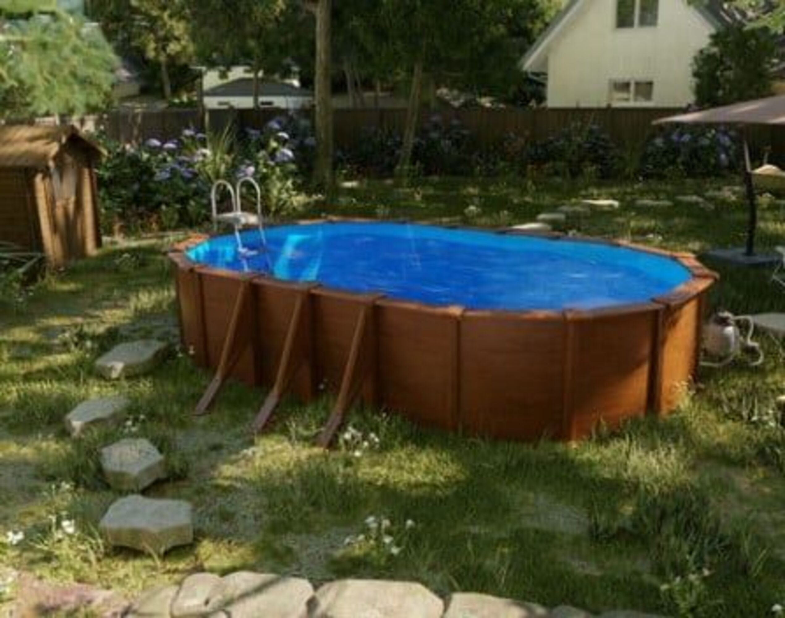 Una piscina elevada con revestimiento de madera se encuentra en un jardín junto a una caseta para perros.
