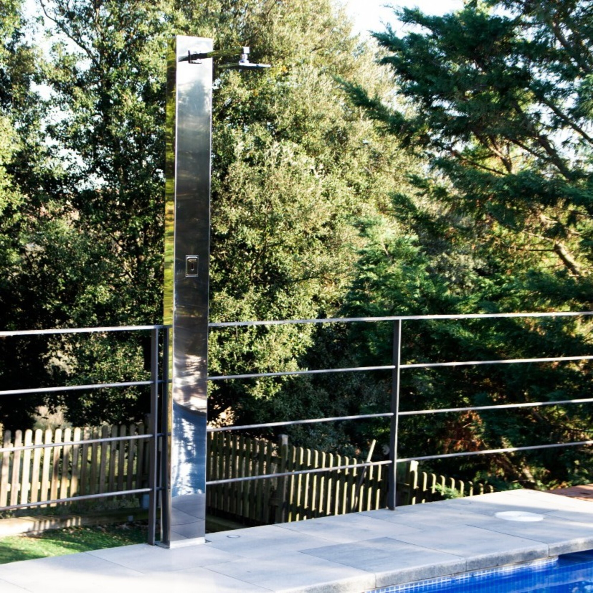 Un poste de ducha brillante junto a una piscina, con árboles al fondo.