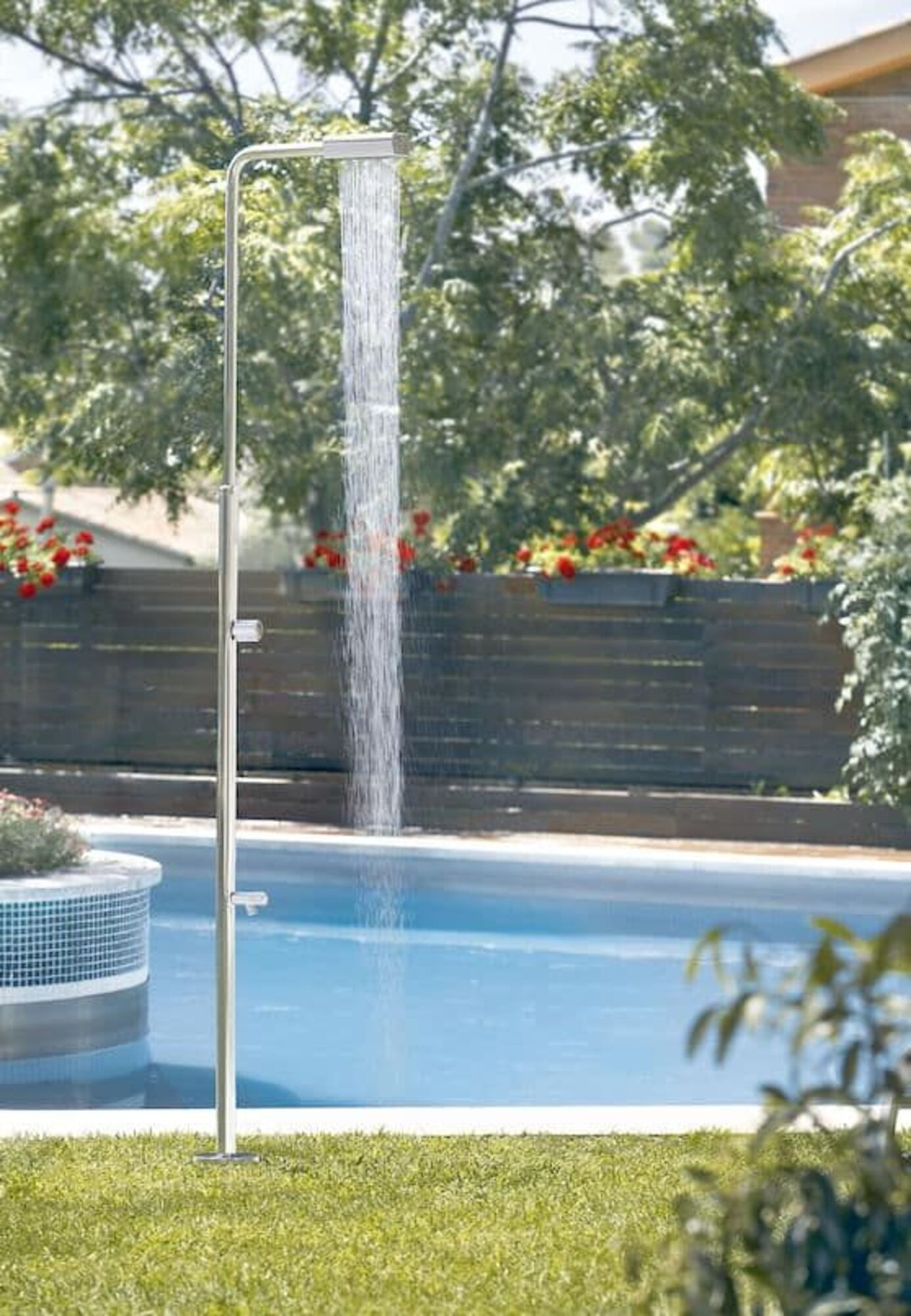 Una ducha exterior con agua corriente junto a una piscina, con árboles y arbustos.