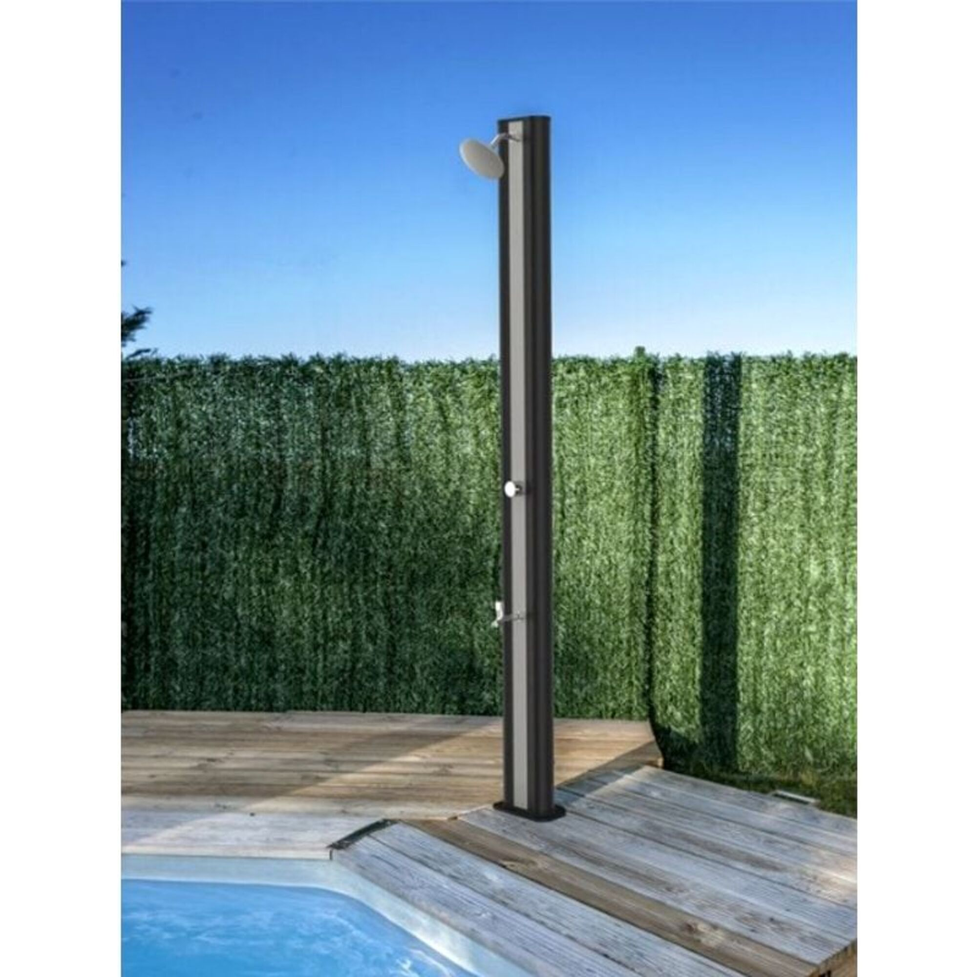 Una columna de ducha oscura con cabezal y grifos en una terraza de madera junto a una piscina.
