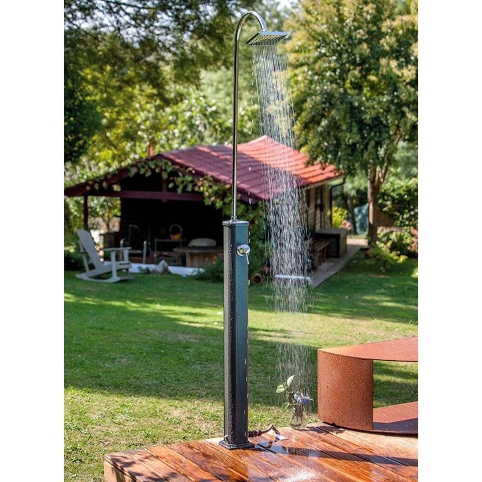 Ducha exterior de jardín con chorro de agua sobre tarima de madera, casa y árboles detrás.
