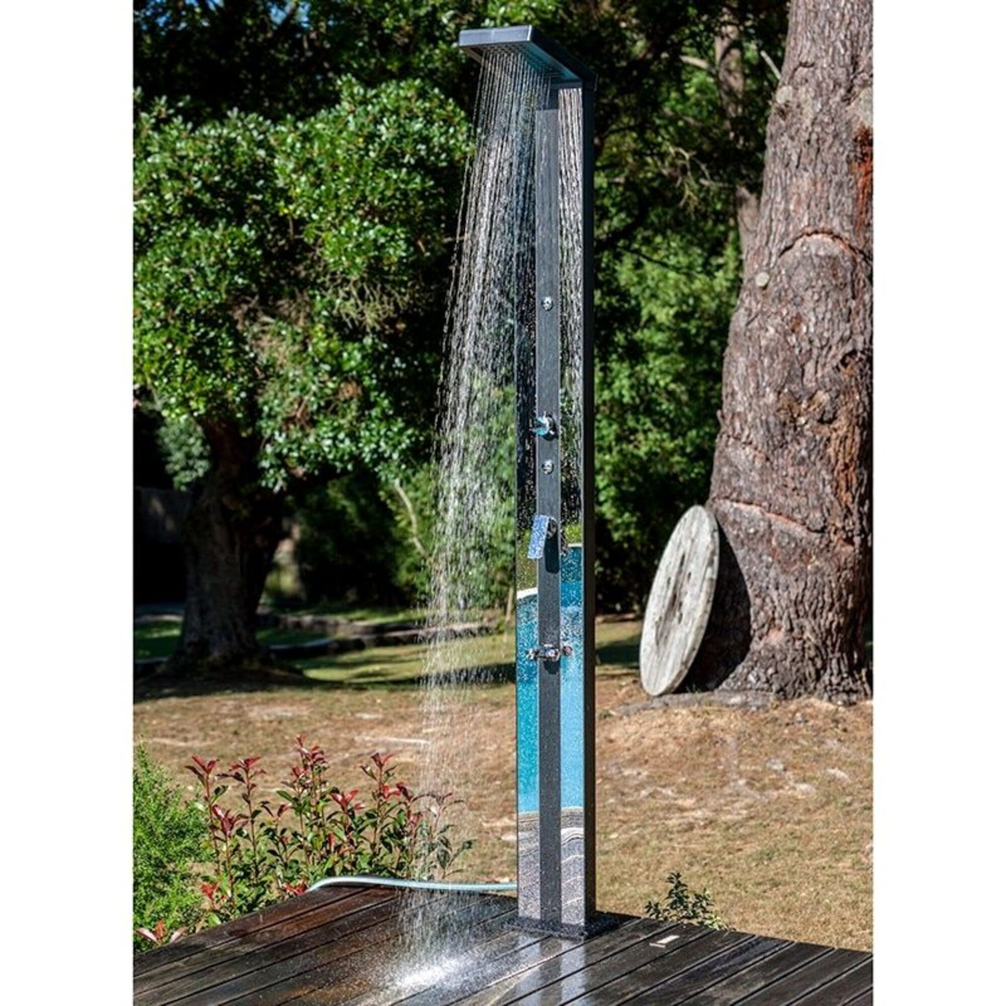 Una ducha exterior moderna con agua fluyendo sobre una terraza de madera, rodeada de árboles.