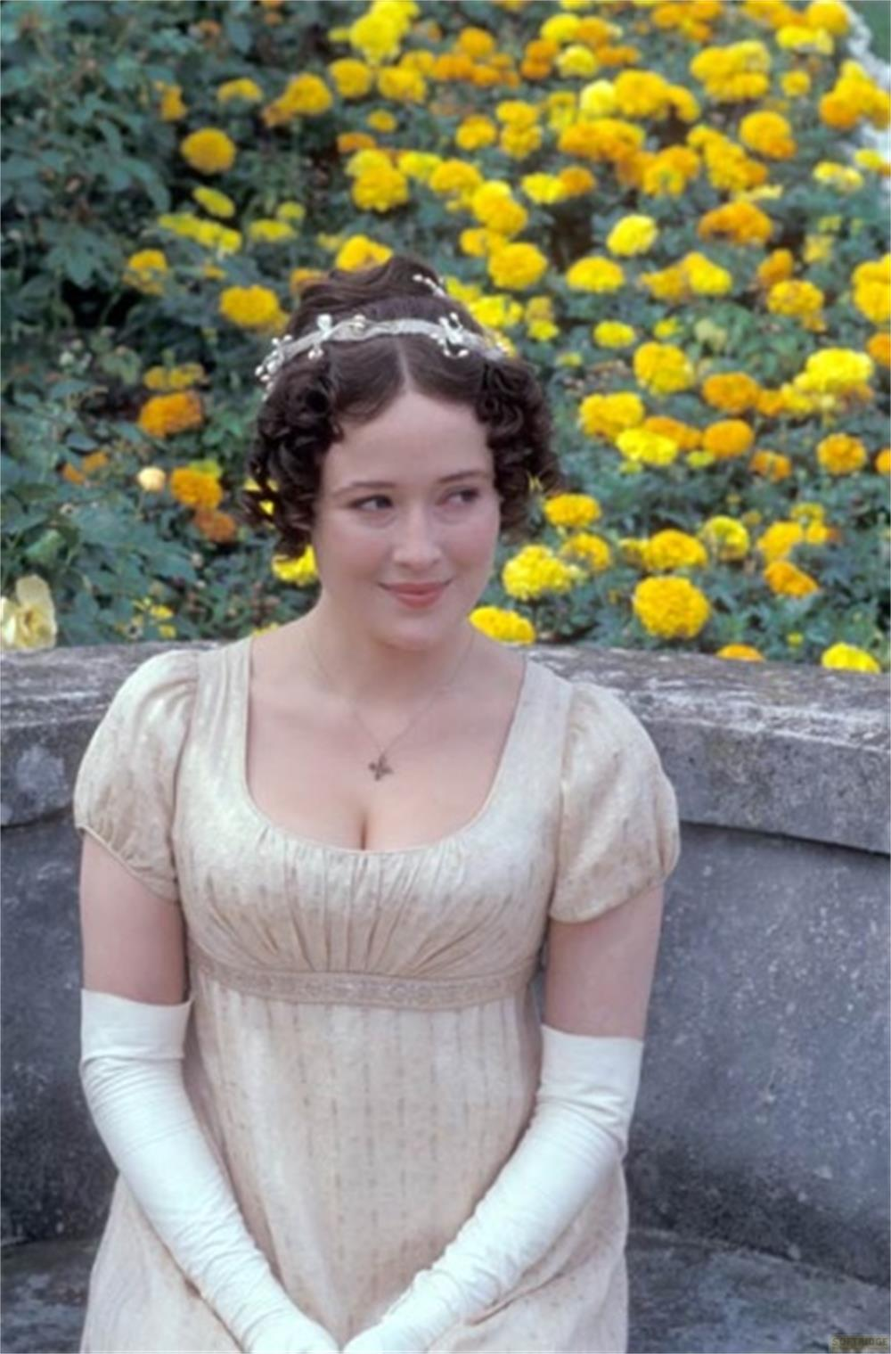 Une femme en robe crème et gants est assise devant des fleurs jaunes.