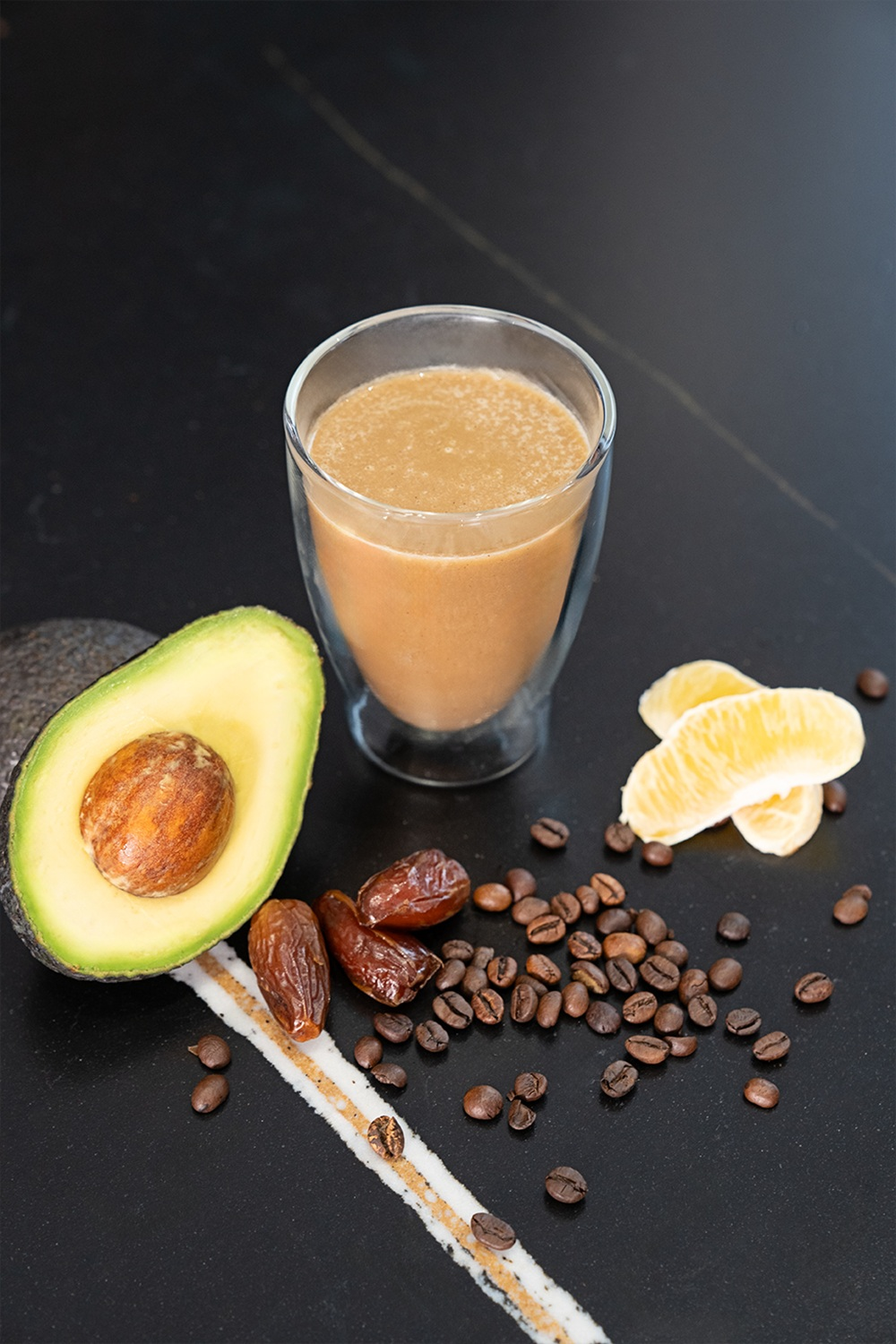 Ein Glas cremiges Getränk neben Avocado, Datteln, Kaffee und Orangenspalten.