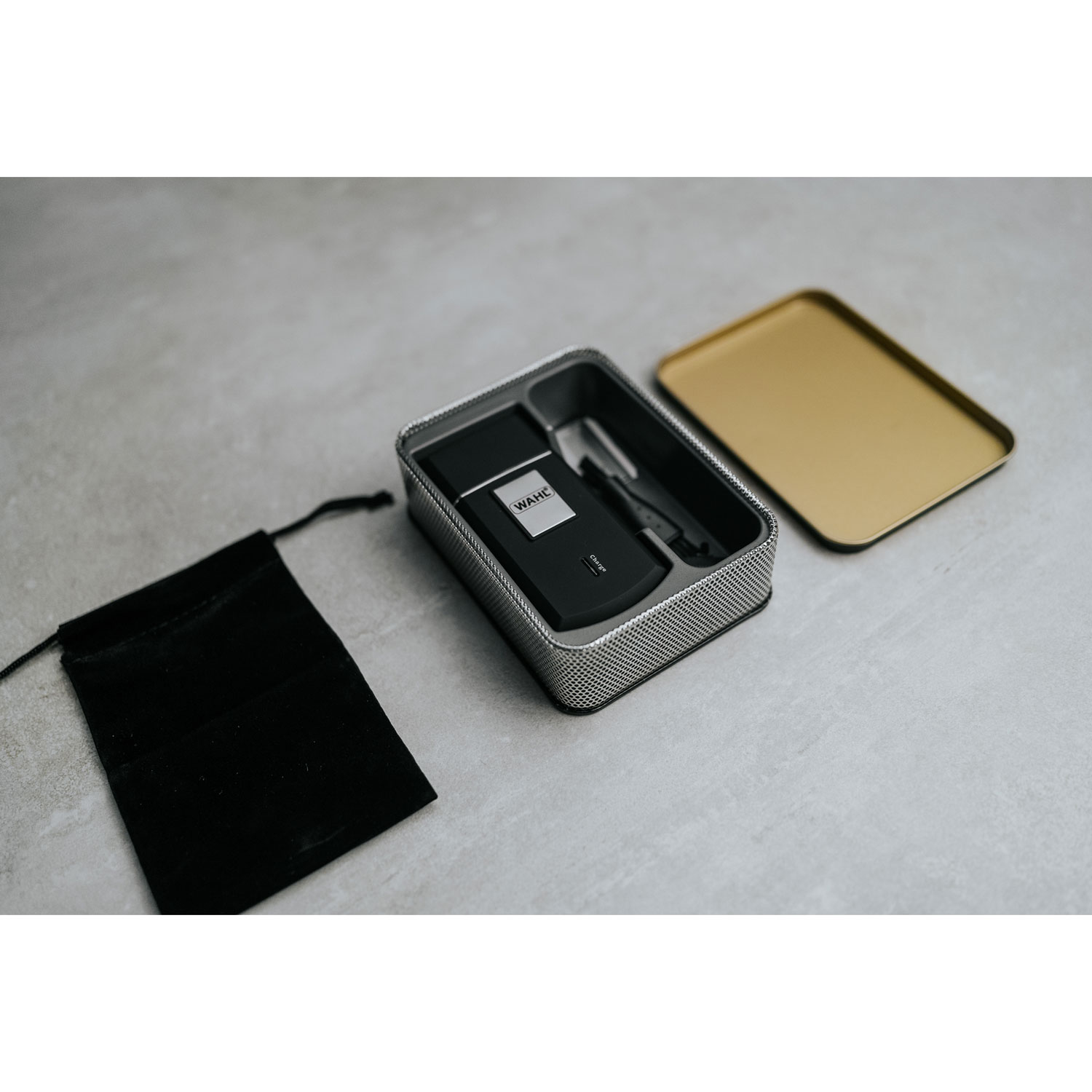 Ein WAHL-Rasierer in einem Metallbehälter, daneben eine schwarze Tasche und ein goldener Deckel.