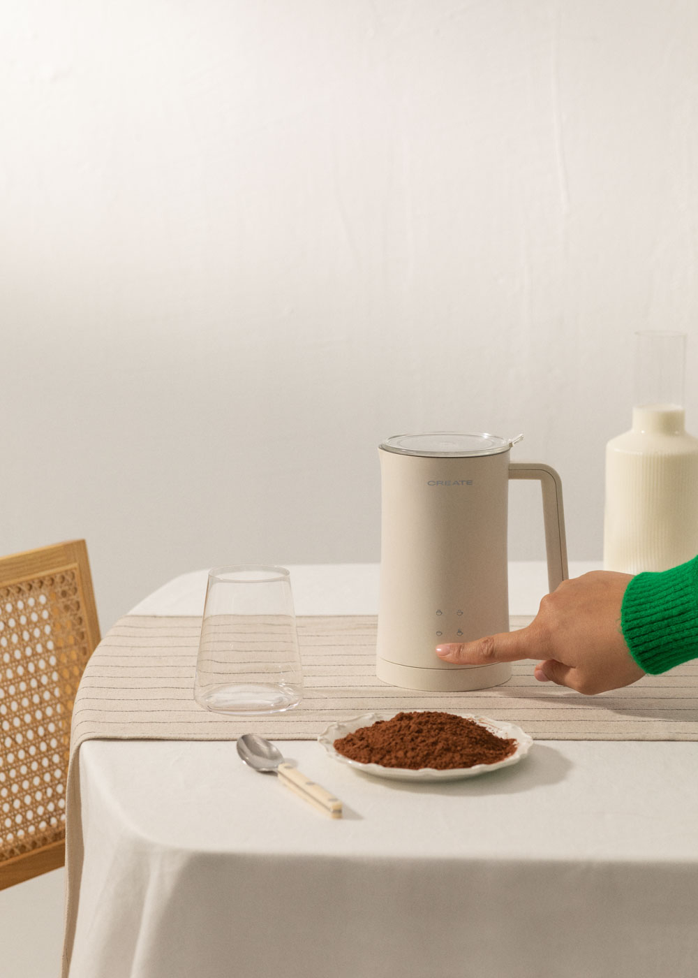 Un dedo presiona botones en un espumador de leche beige. Cacao en polvo, cuchara y vaso en mesa.