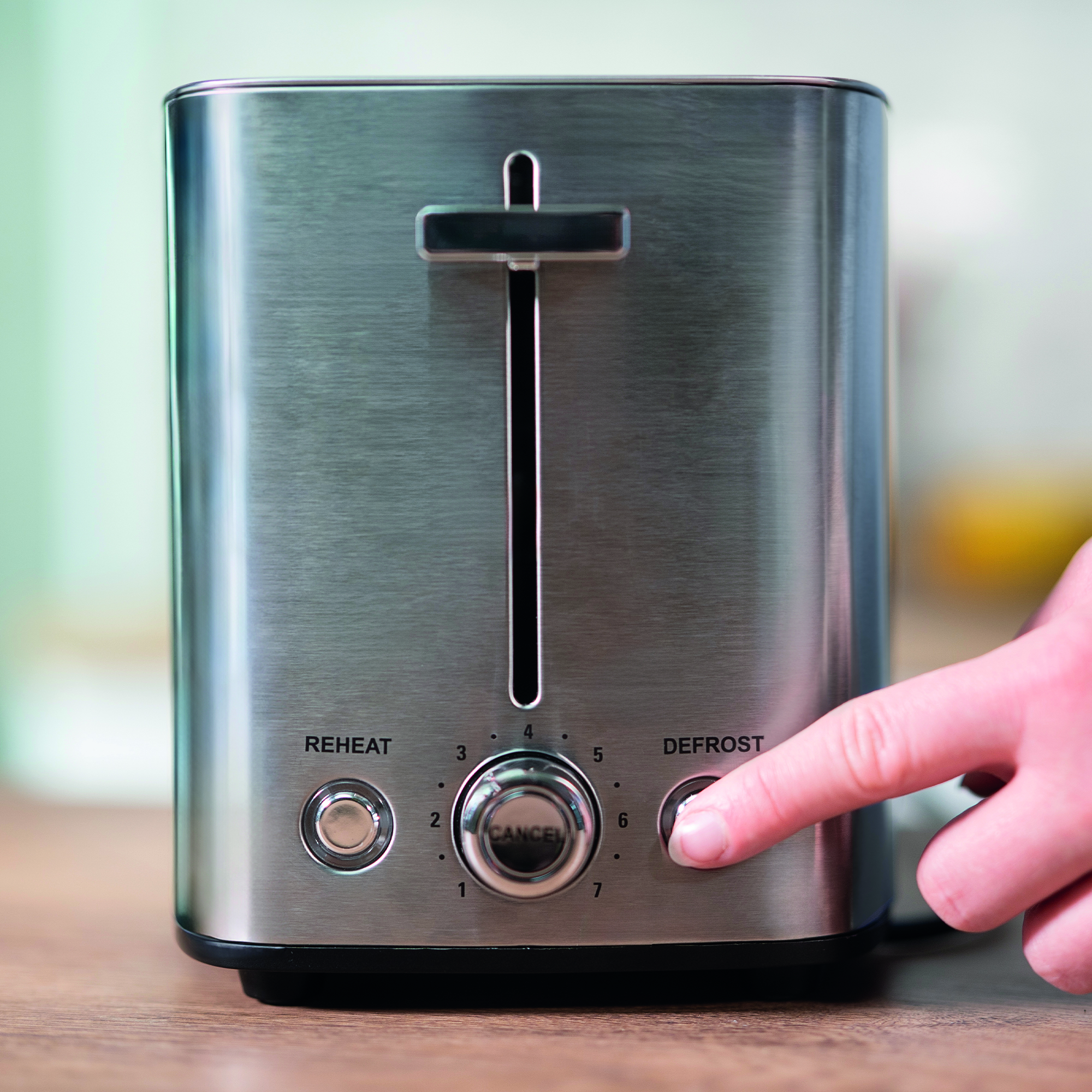 Ein Finger drückt die Defrost-Taste eines silbernen Toasters auf einem Holztisch.