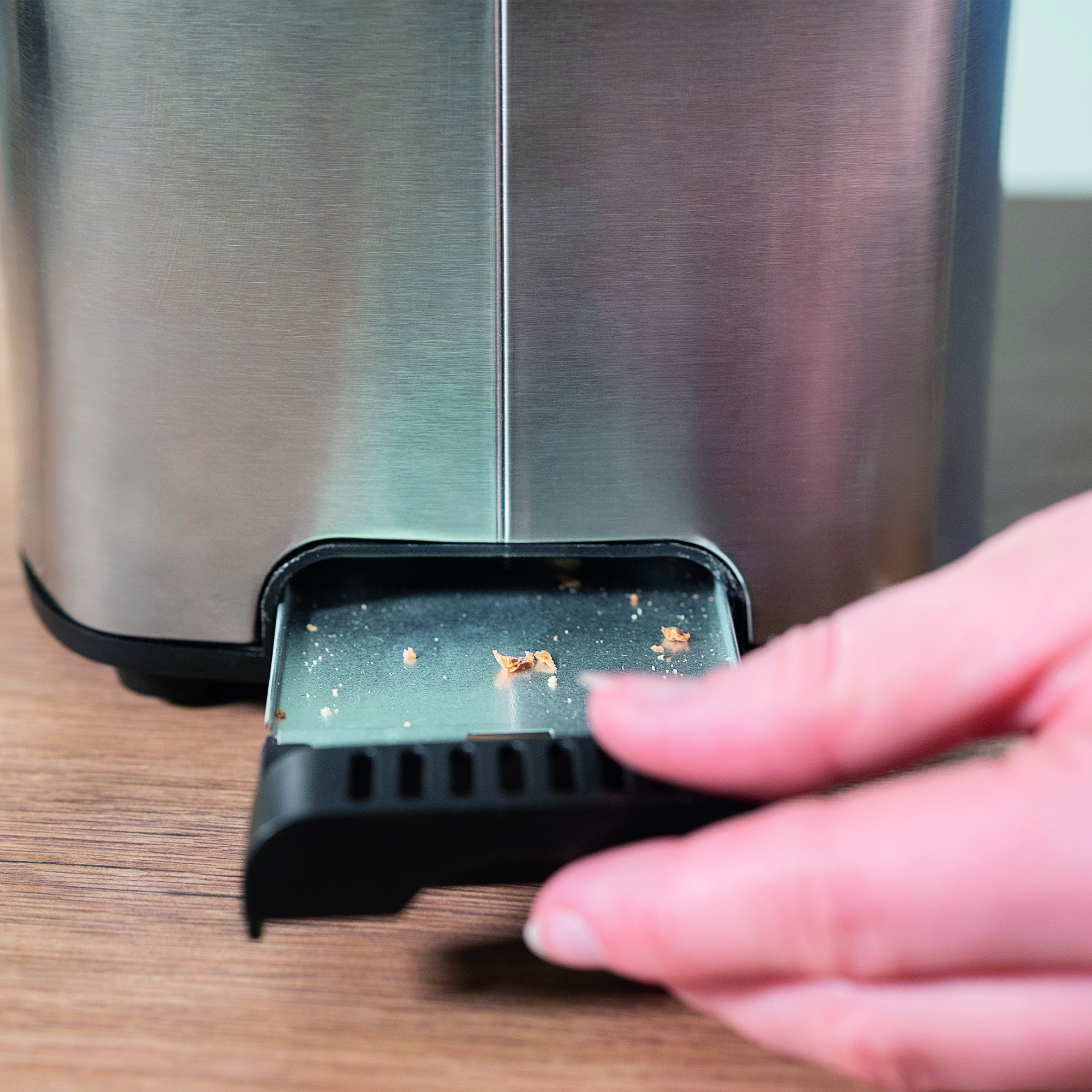 Eine Hand zieht die Krümelschublade eines Toasters heraus. Kleine Brotkrümel sind sichtbar.