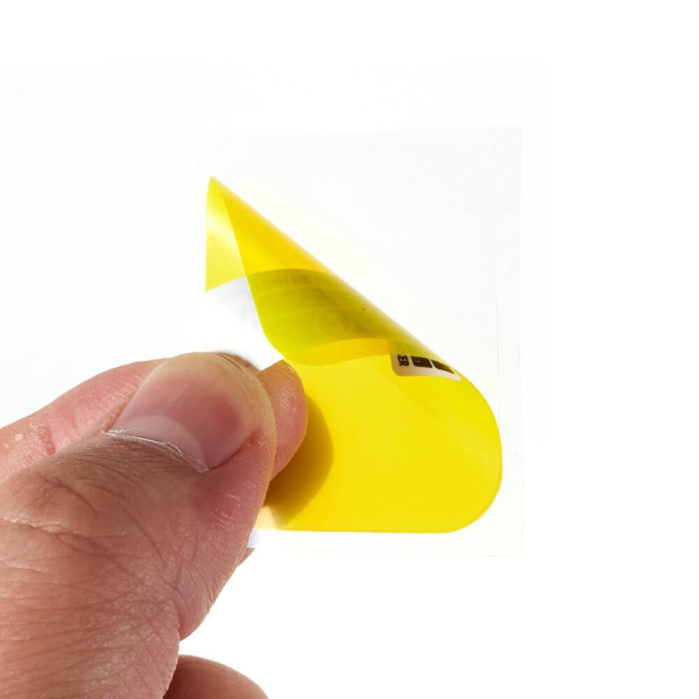 Une main tient un autocollant jaune qui se courbe. L'autocollant porte des marques noires.