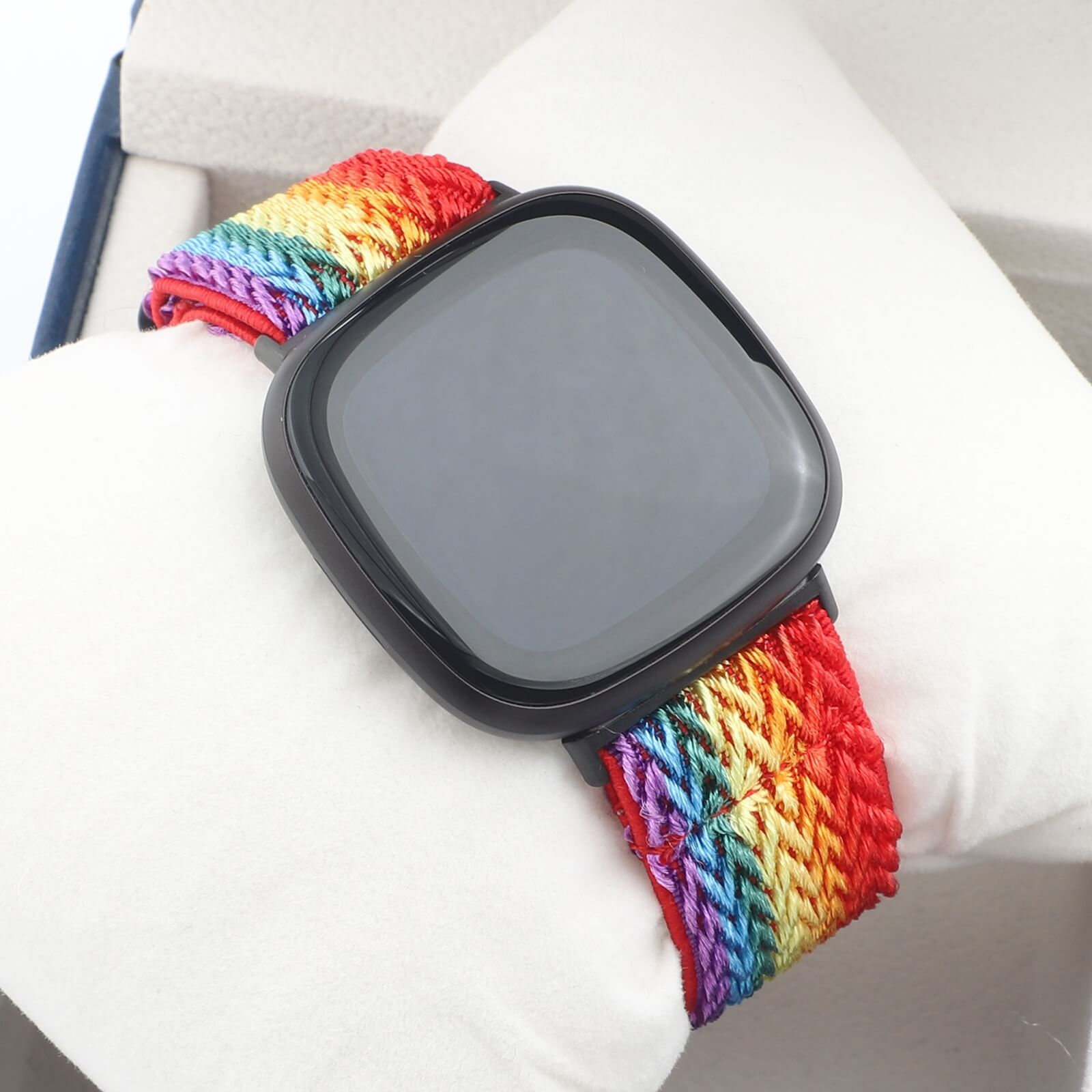 Une montre connectée noire avec bracelet arc-en-ciel repose sur un coussin blanc.