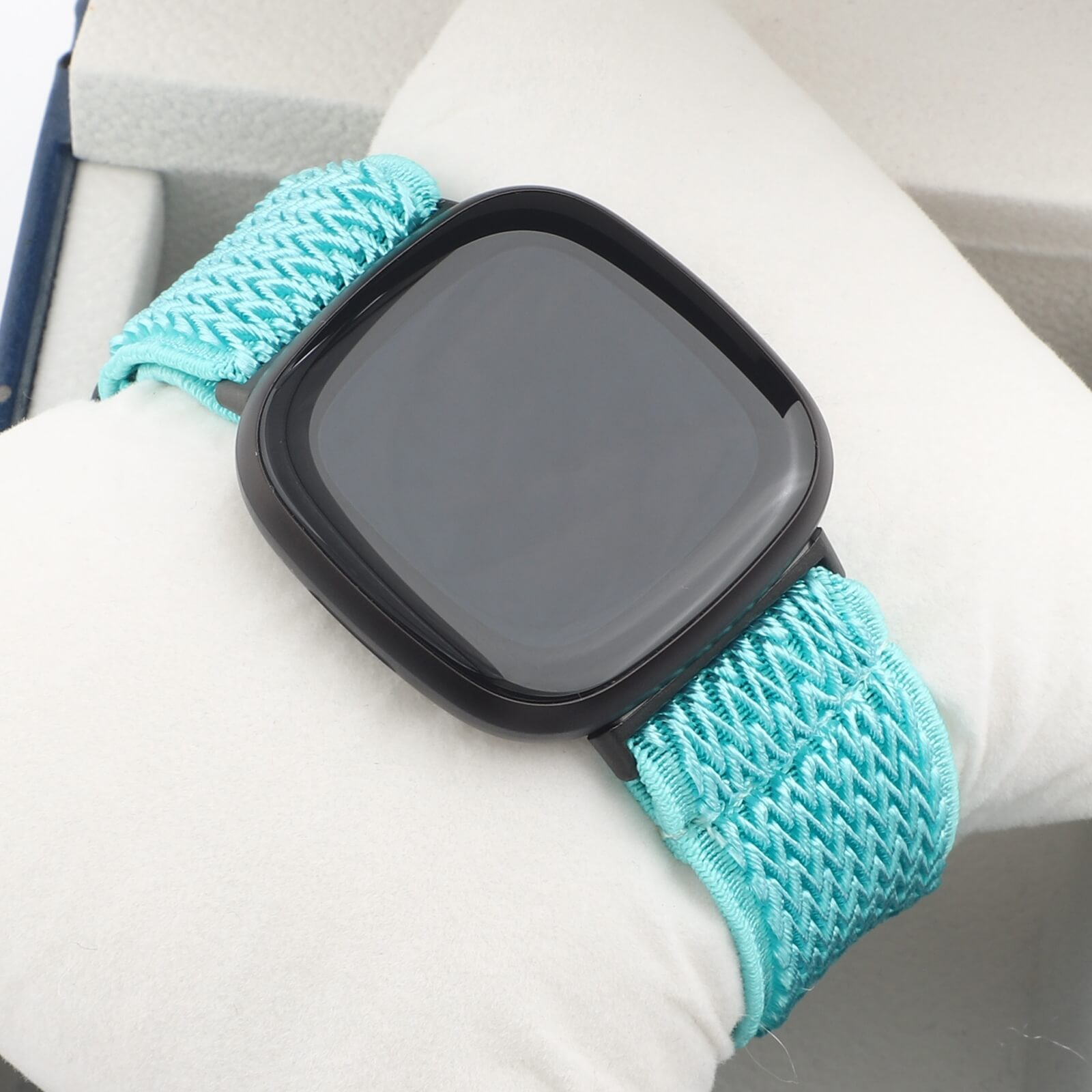 Montre connectée noire avec bracelet turquoise sur coussin blanc.