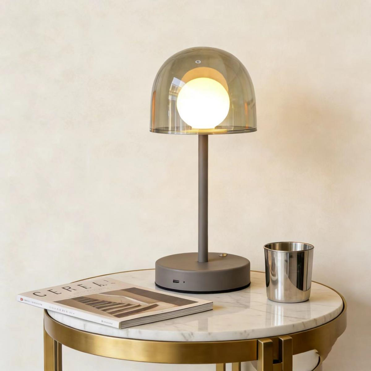 Graue Lampe mit Glaskuppel auf Marmortisch neben Buch und Becher.