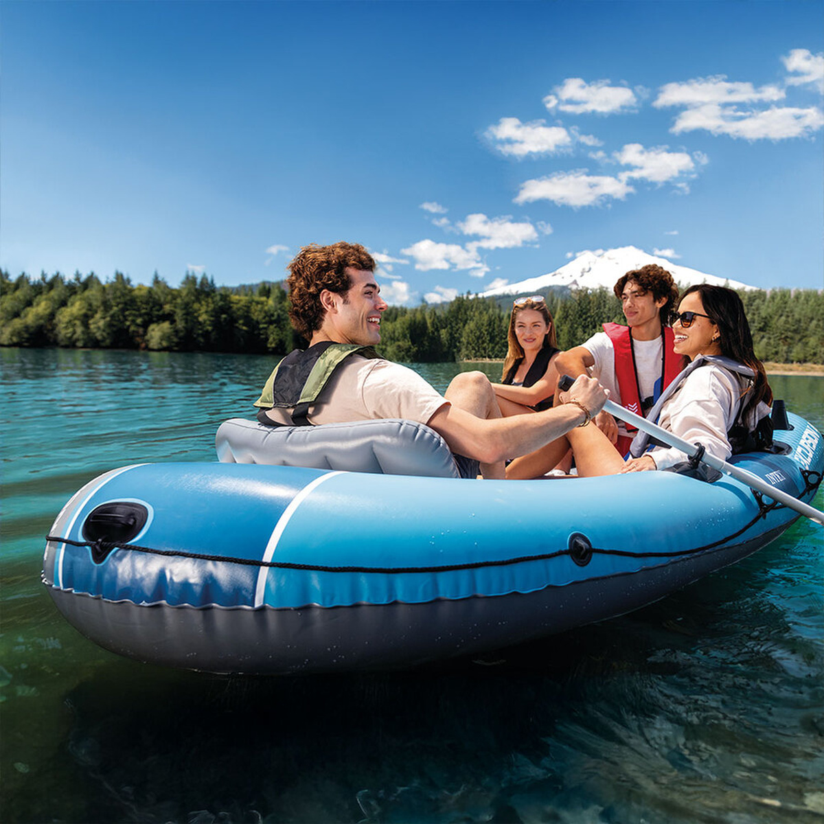 Vier Personen in einem blauen Schlauchboot auf einem See mit einem schneebedeckten Berg im Hintergrund.