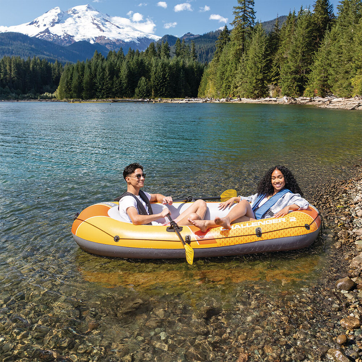 Zwei Personen in einem orangefarbenen Schlauchboot auf einem klaren See, mit schneebedecktem Berg und Wald.