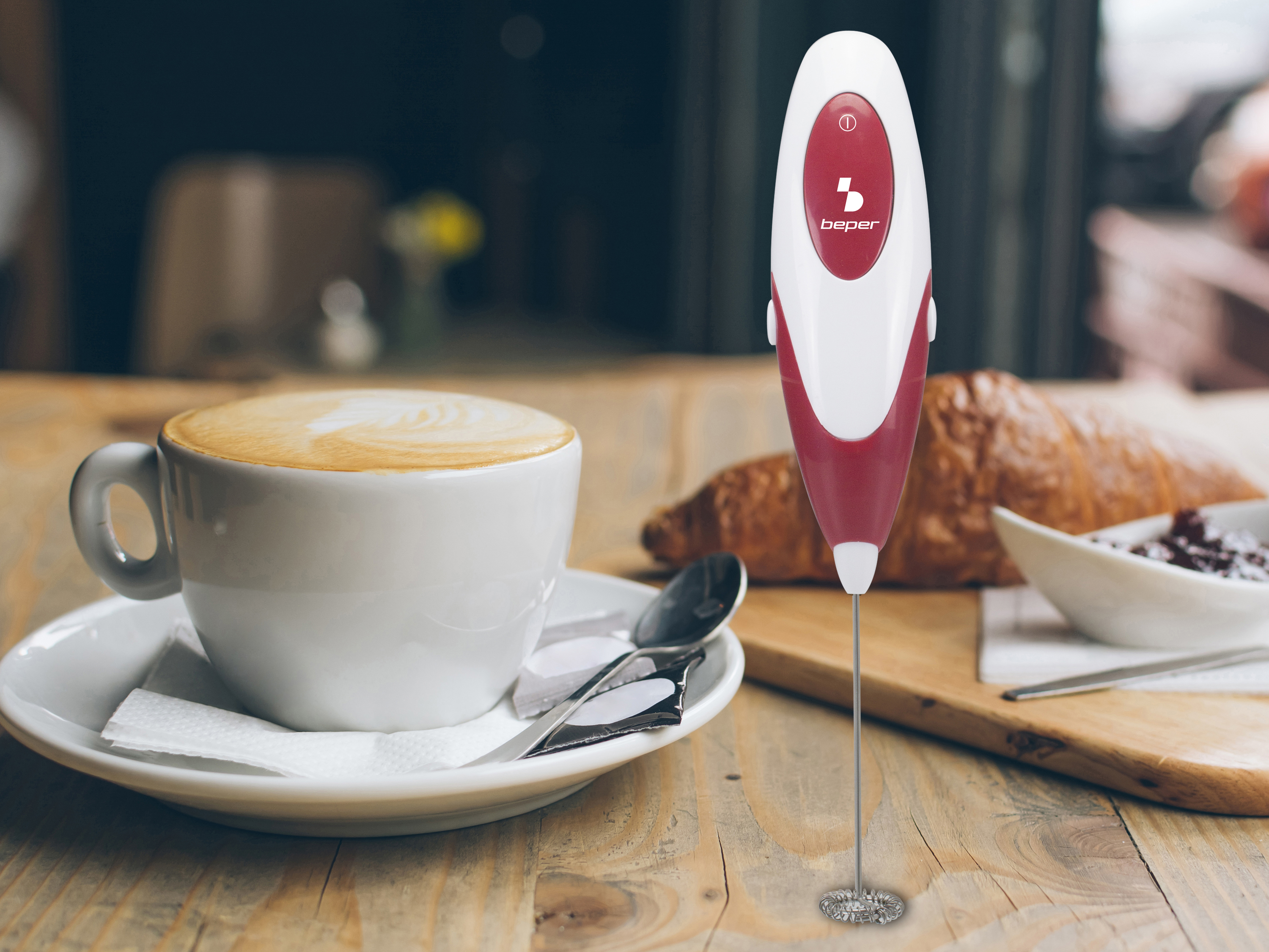 Sbattitore per latte accanto a cappuccino e croissant su tavolo di legno.