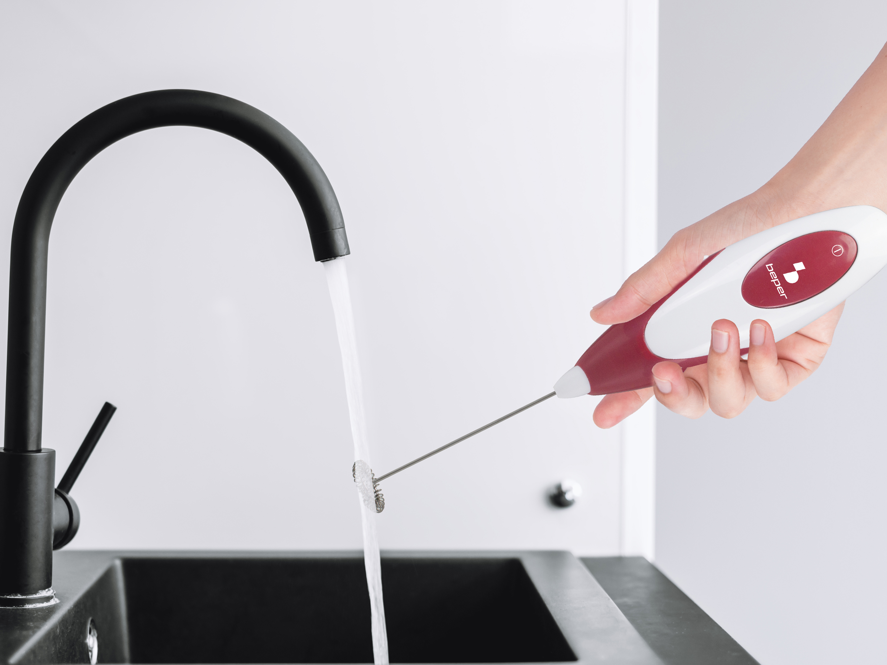 Una mano tiene un montalatte bianco e rosso sopra un lavello nero con acqua corrente.