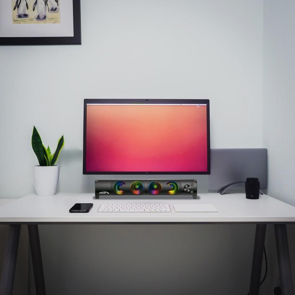 Un escritorio con monitor, teclado, altavoz y planta.