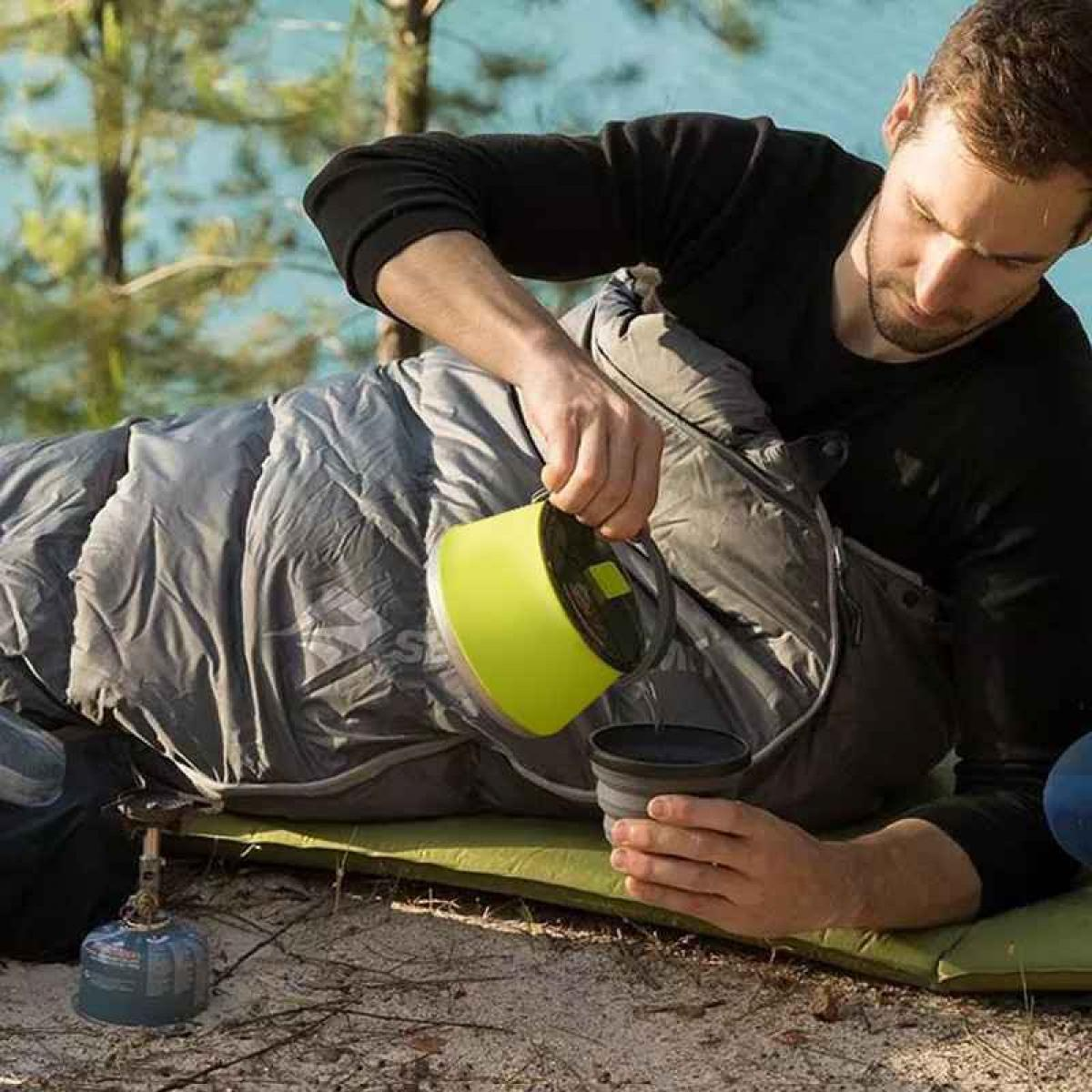 Mann gießt aus gelber Kanne in graue Tasse, liegt im Schlafsack.