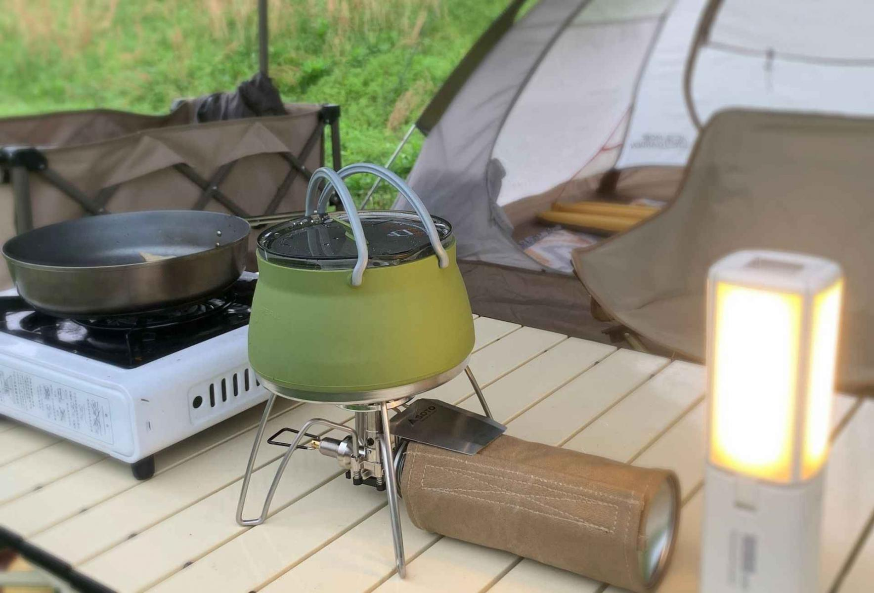 Campingkocher mit Topf, Pfanne und Zelt im Hintergrund.