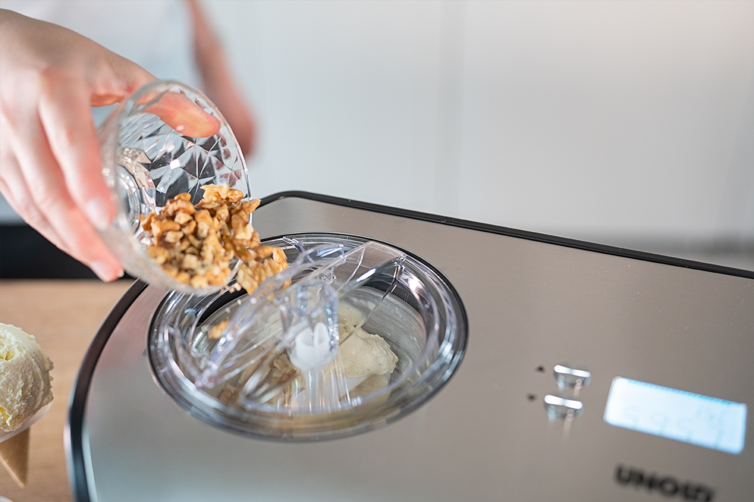 Una mano vierte nueces picadas en una máquina de helados con tapa transparente.
