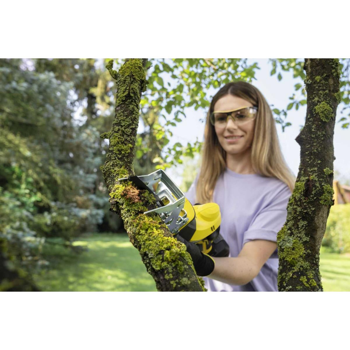 Frau mit Schutzbrille schneidet Baum mit gelber Säge auf moosbewachsenem Ast.