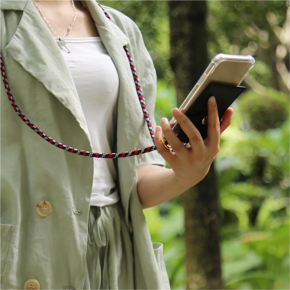Persona che tiene telefono con cordino. Giacca verde, camicia bianca, sfondo natura.