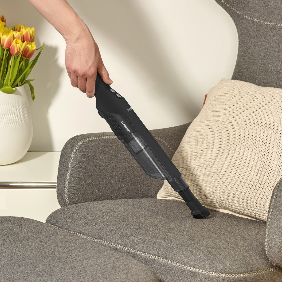 Main passant l'aspirateur sur un canapé gris avec un aspirateur noir.