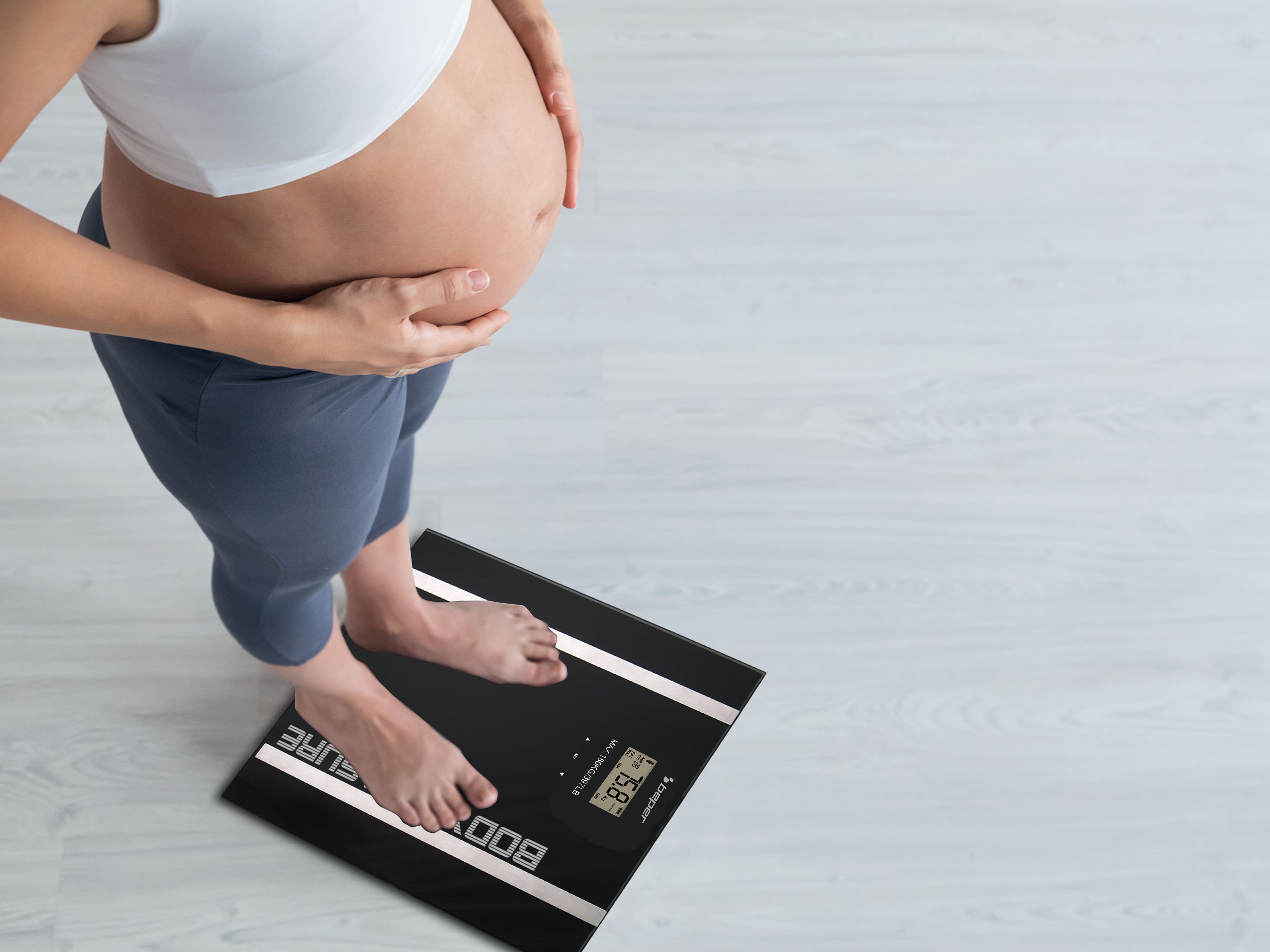 Kobieta w ciąży na wadze. Waga 75,8 kg. Jasna drewniana podłoga.