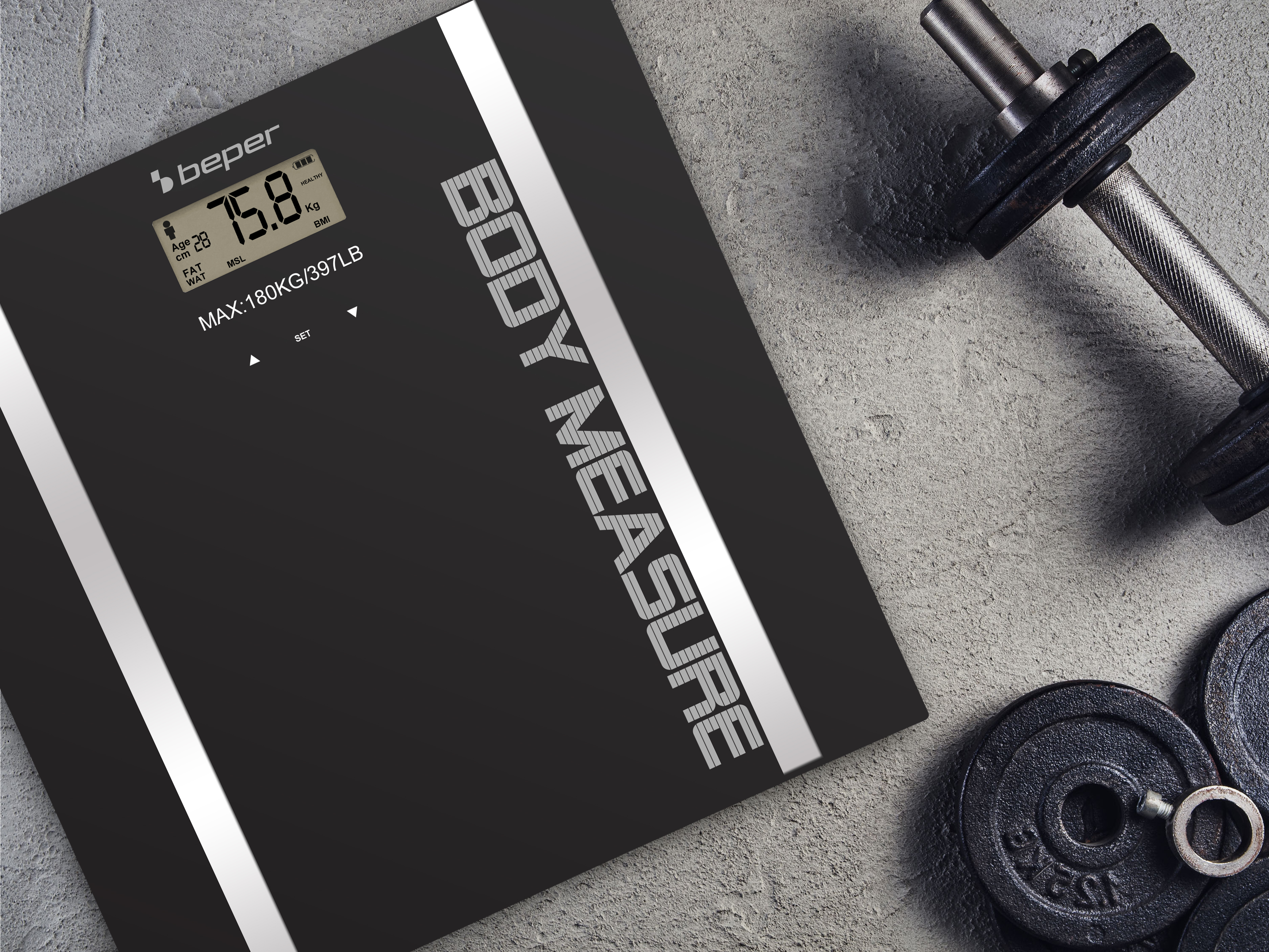 Czarna cyfrowa waga łazienkowa pokazuje 75,8 kg. W pobliżu znajdują się hantle i talerze obciążeniowe.