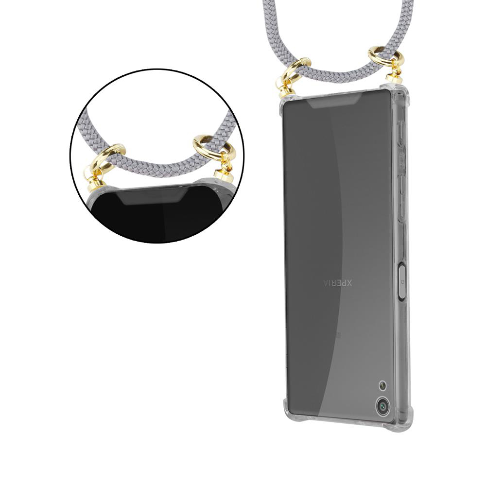 Graues Handy-Lanyard mit goldenen Ringen an einem transparenten Kasten.