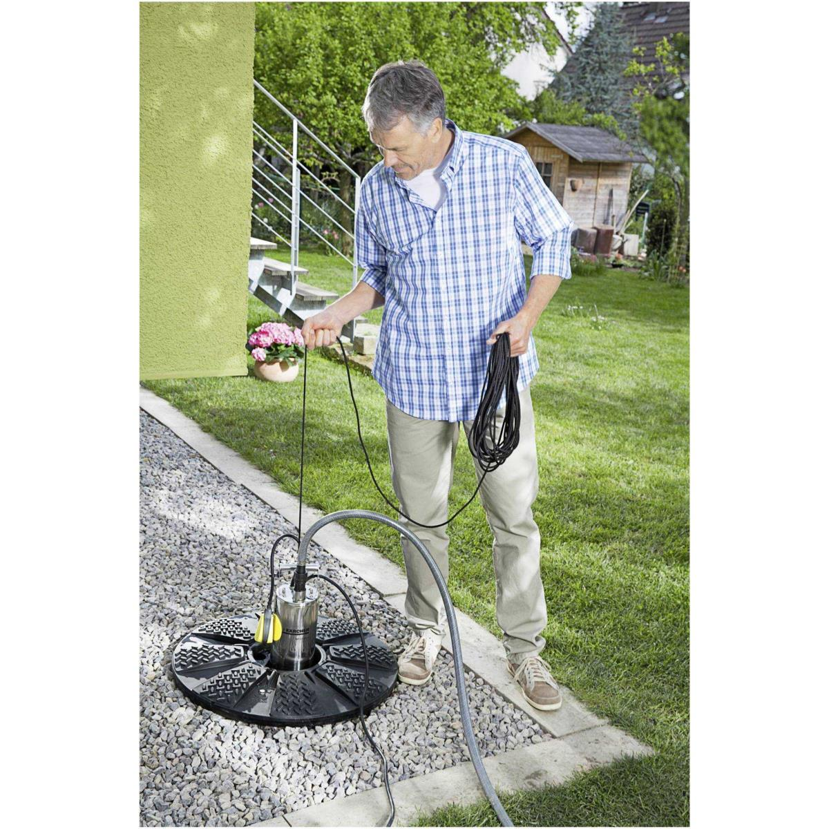 Mann mit blauer karierter Hemd hält Kabel neben Tauchpumpe im Garten.