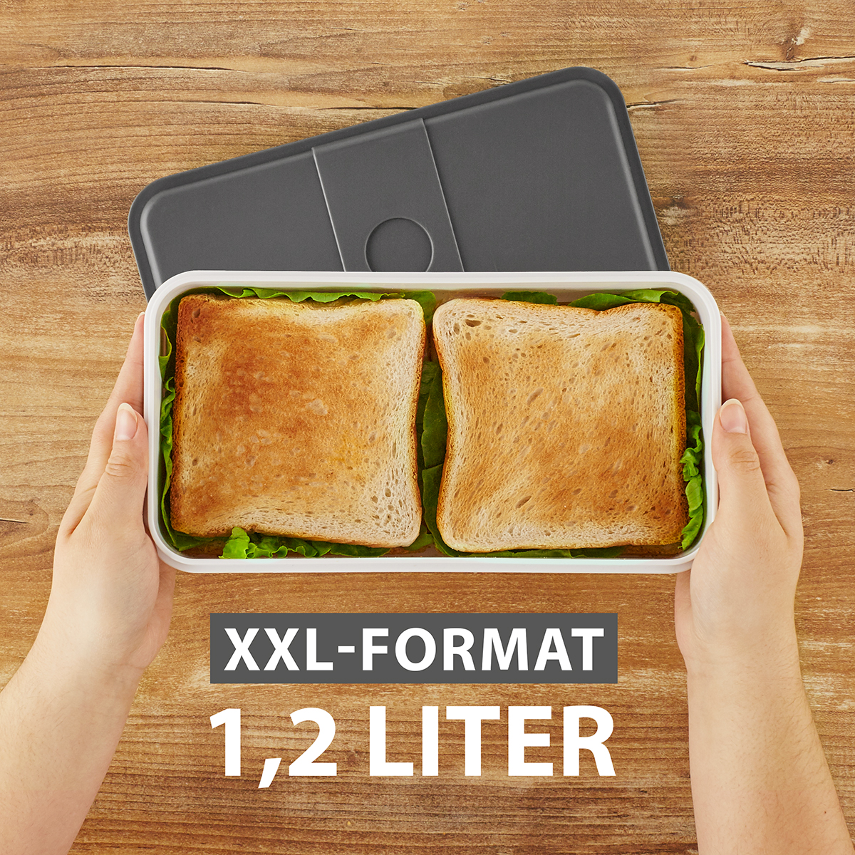 Hände halten eine Brotdose mit zwei Sandwiches und Salat auf Holzuntergrund.