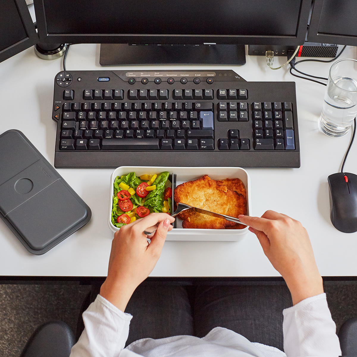 Eine Person isst ein Schnitzel mit Salat am Schreibtisch, daneben Tastatur, Maus und Computer.