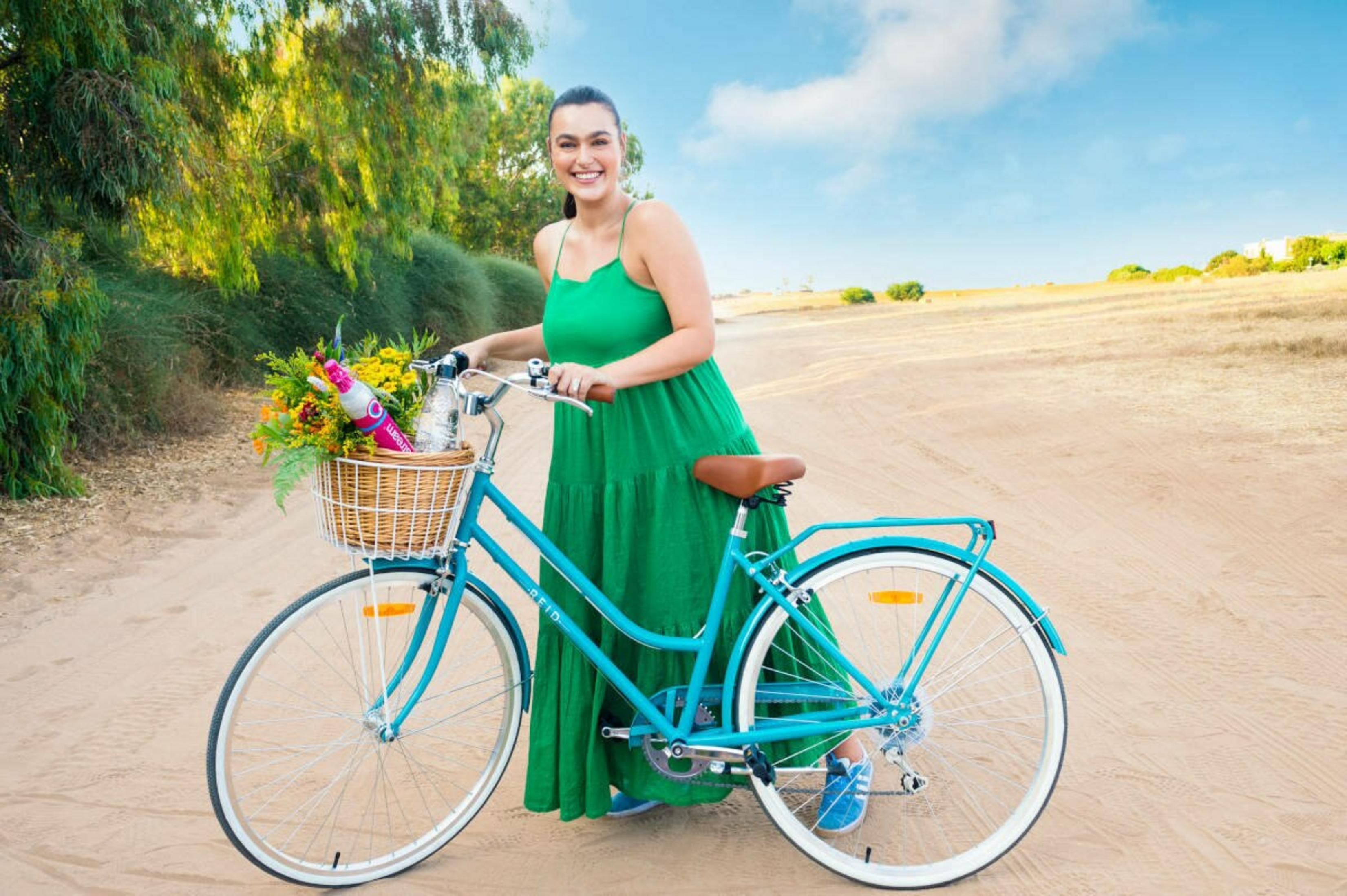 Kobieta z rowerem na polnej drodze, kwiaty i butelka w koszyku.