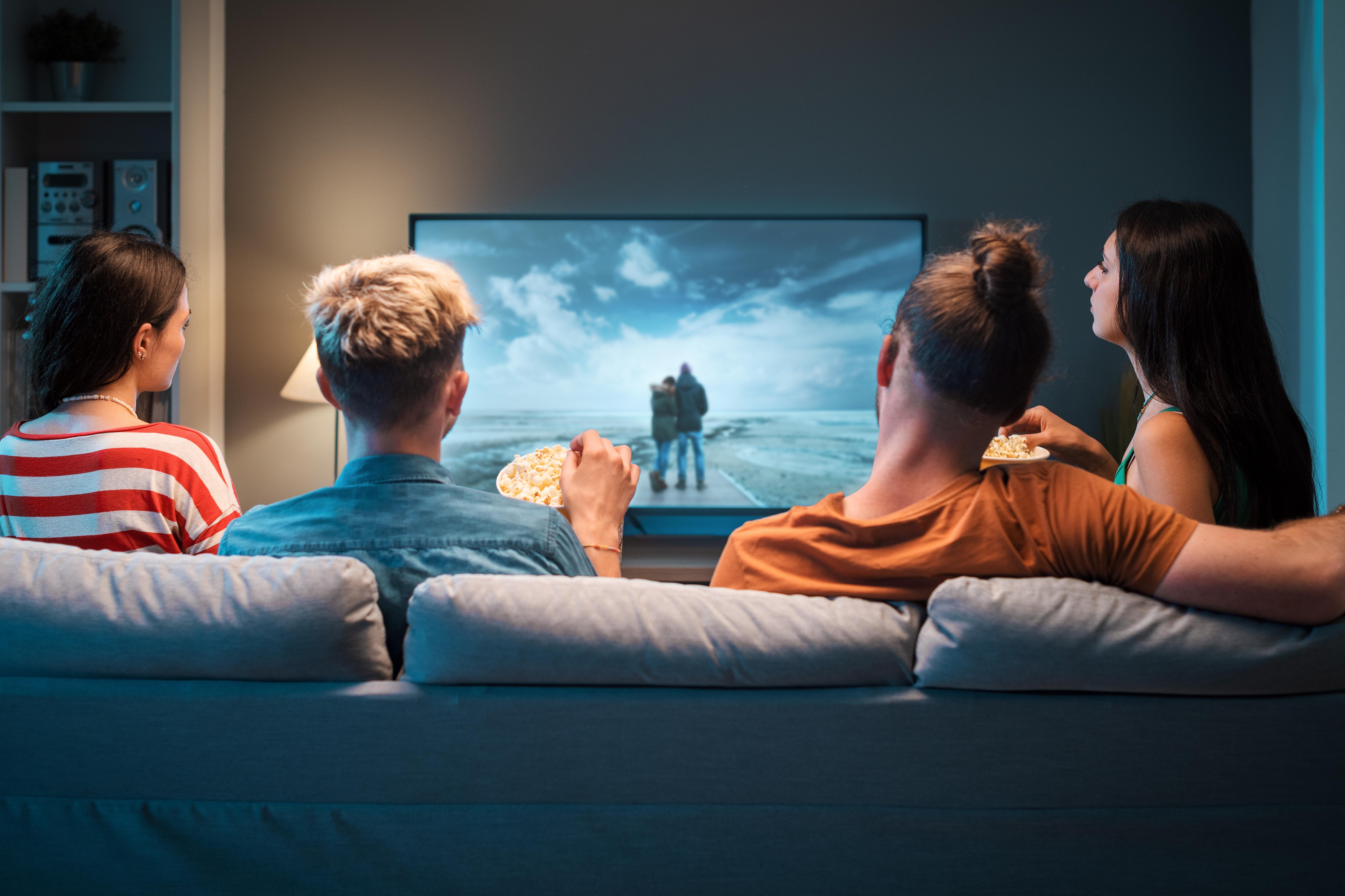 Vier Personen sitzen auf einem Sofa und schauen fern. Einer isst Popcorn.