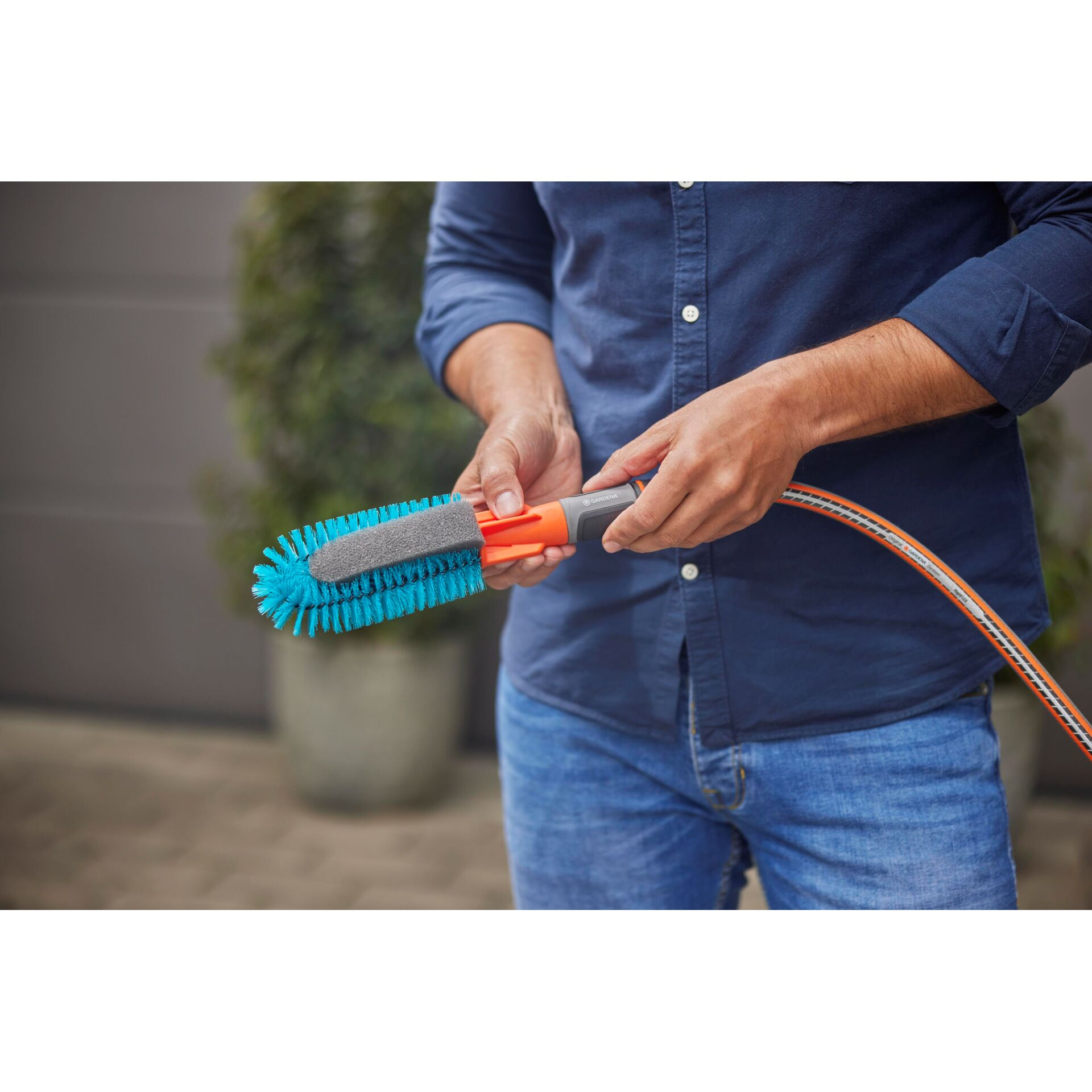 Uomo tiene tubo da giardino e spazzola con setole blu e manico arancione.