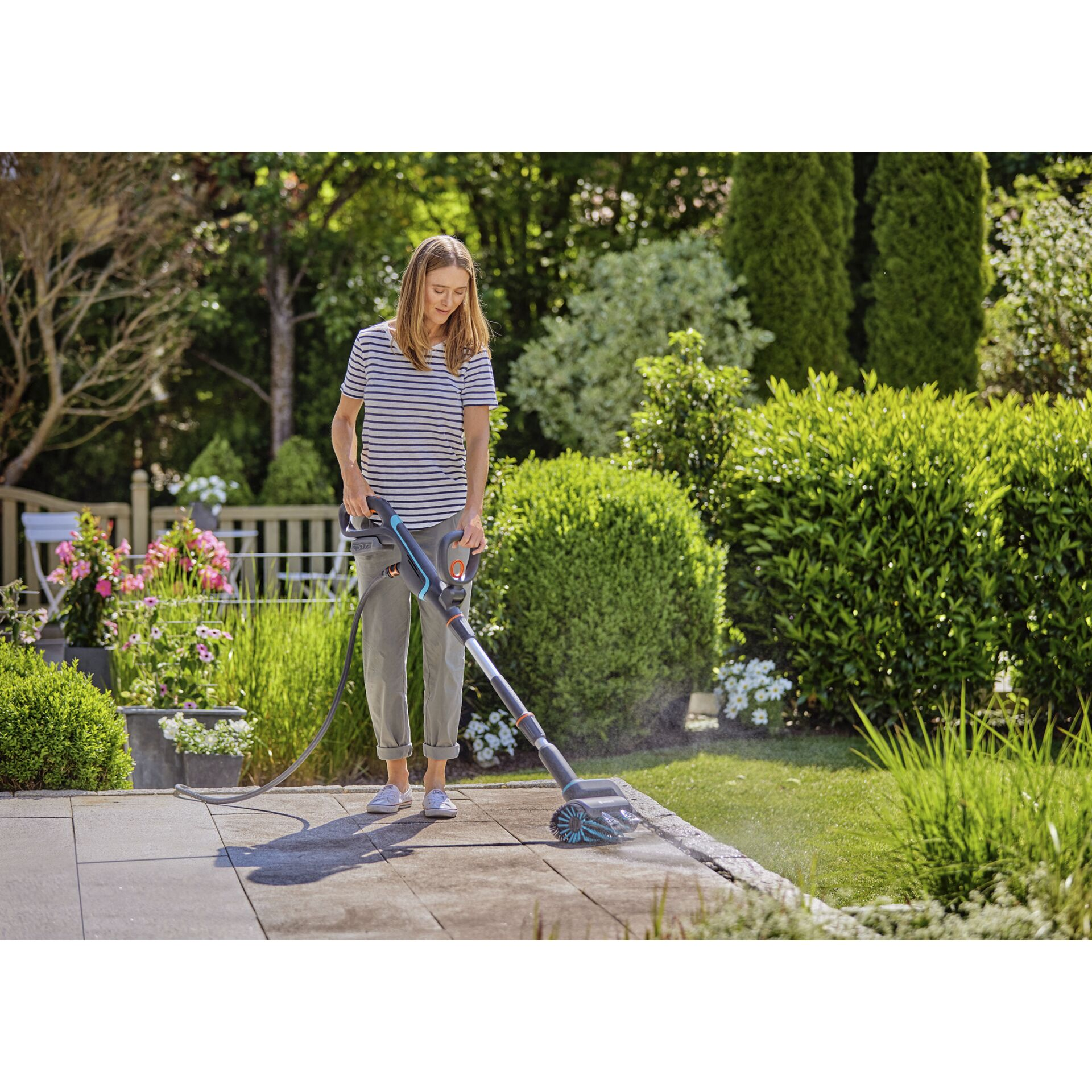 Femme nettoyant une terrasse avec un nettoyeur à brosse dans un jardin verdoyant.
