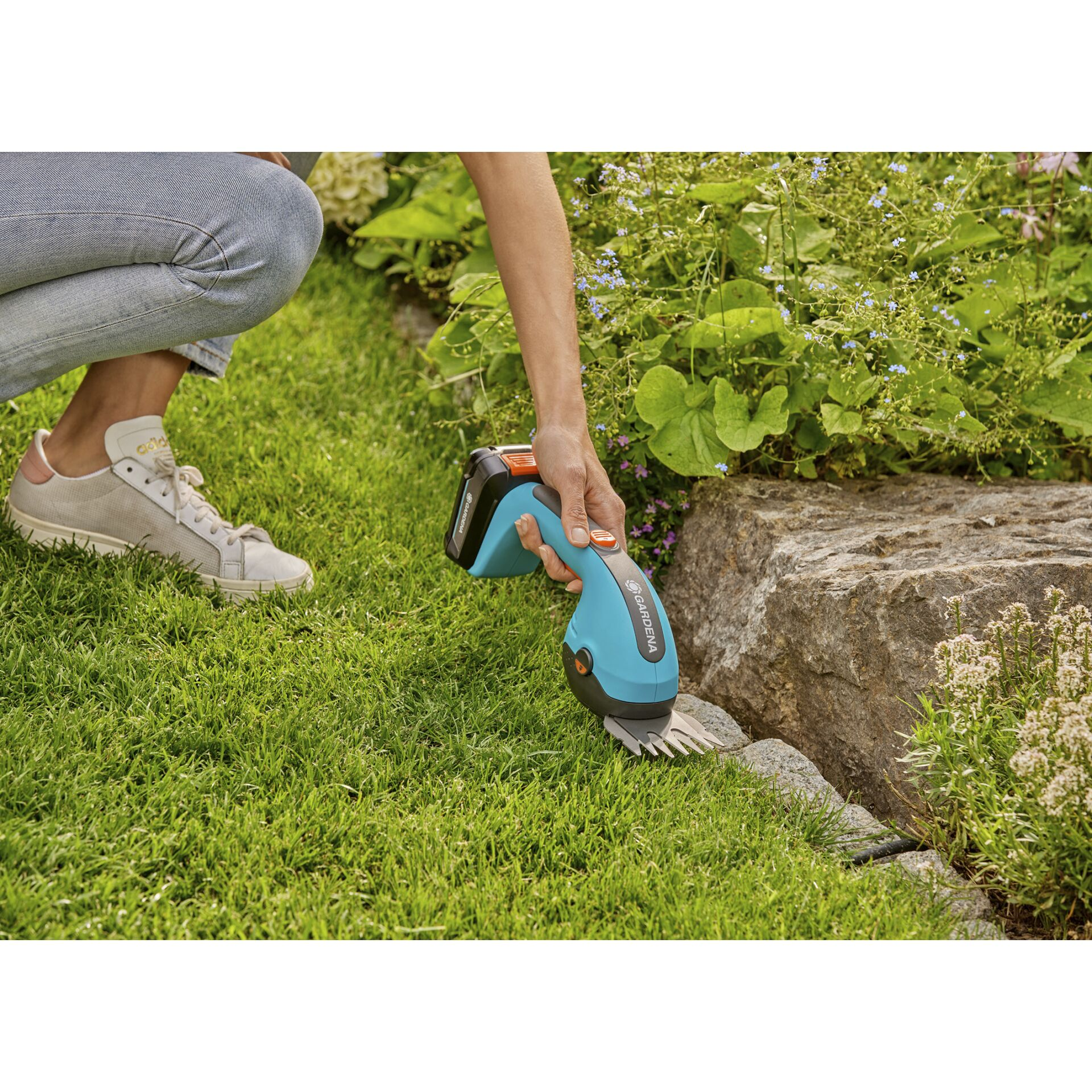 Osoba przycina brzegi trawnika nożycami akumulatorowymi obok kamienia i kwiatów.
