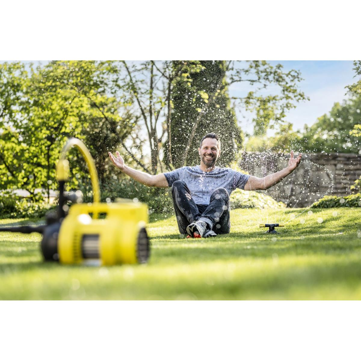Mann sitzt im Gras, bespritzt von Wasser aus einem Sprinkler und einer gelben Maschine.